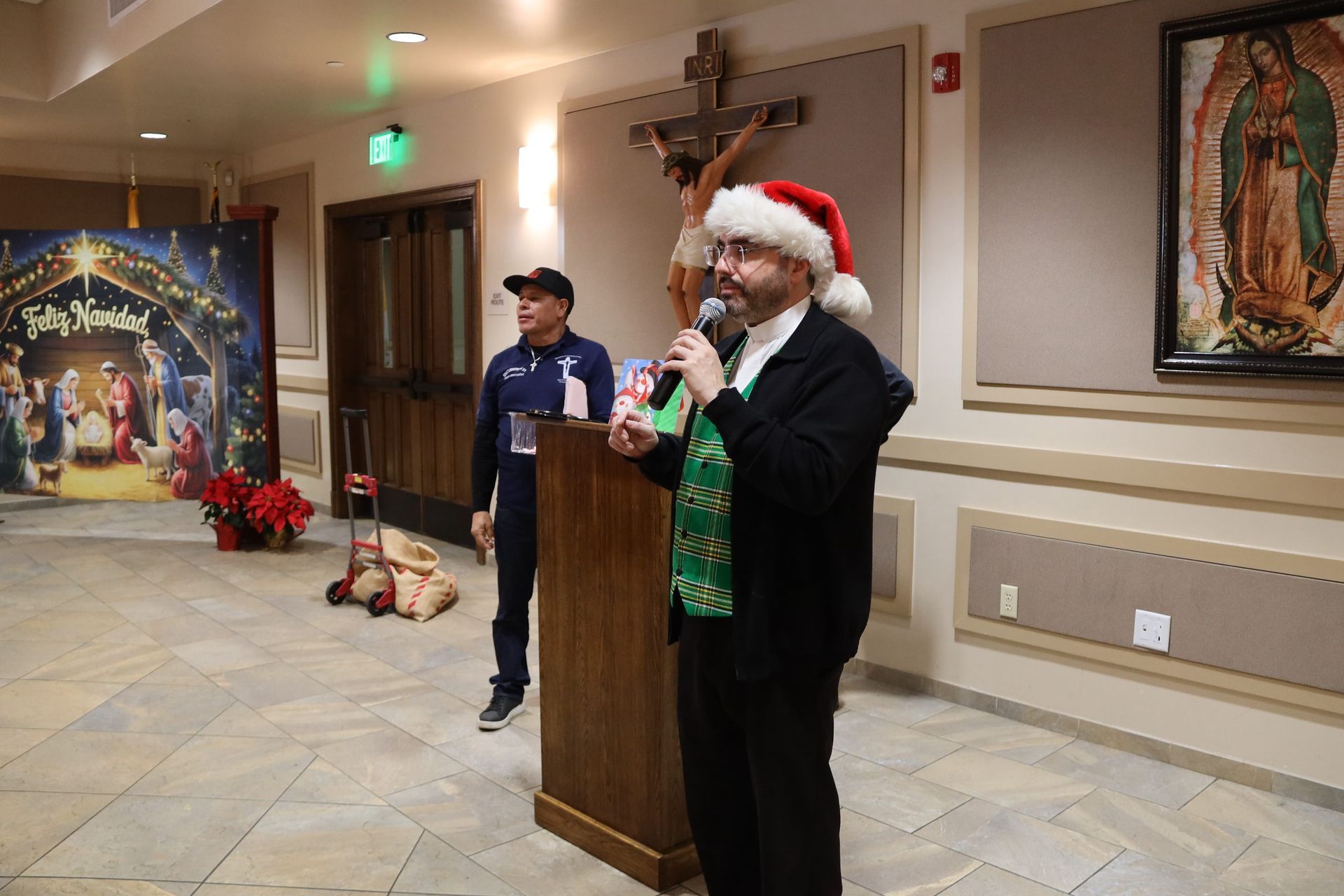Santa-hatted speaker at church podium beside a child, with Christmas decorations and nativity scene.