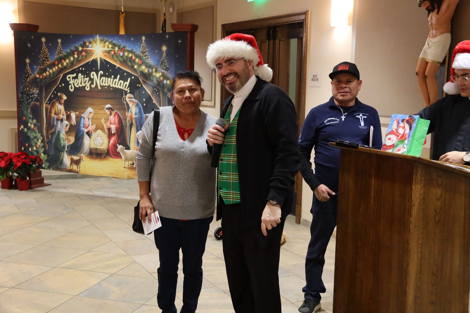 Two people pose indoors in front of a festive holiday backdrop, one wearing a Santa hat.