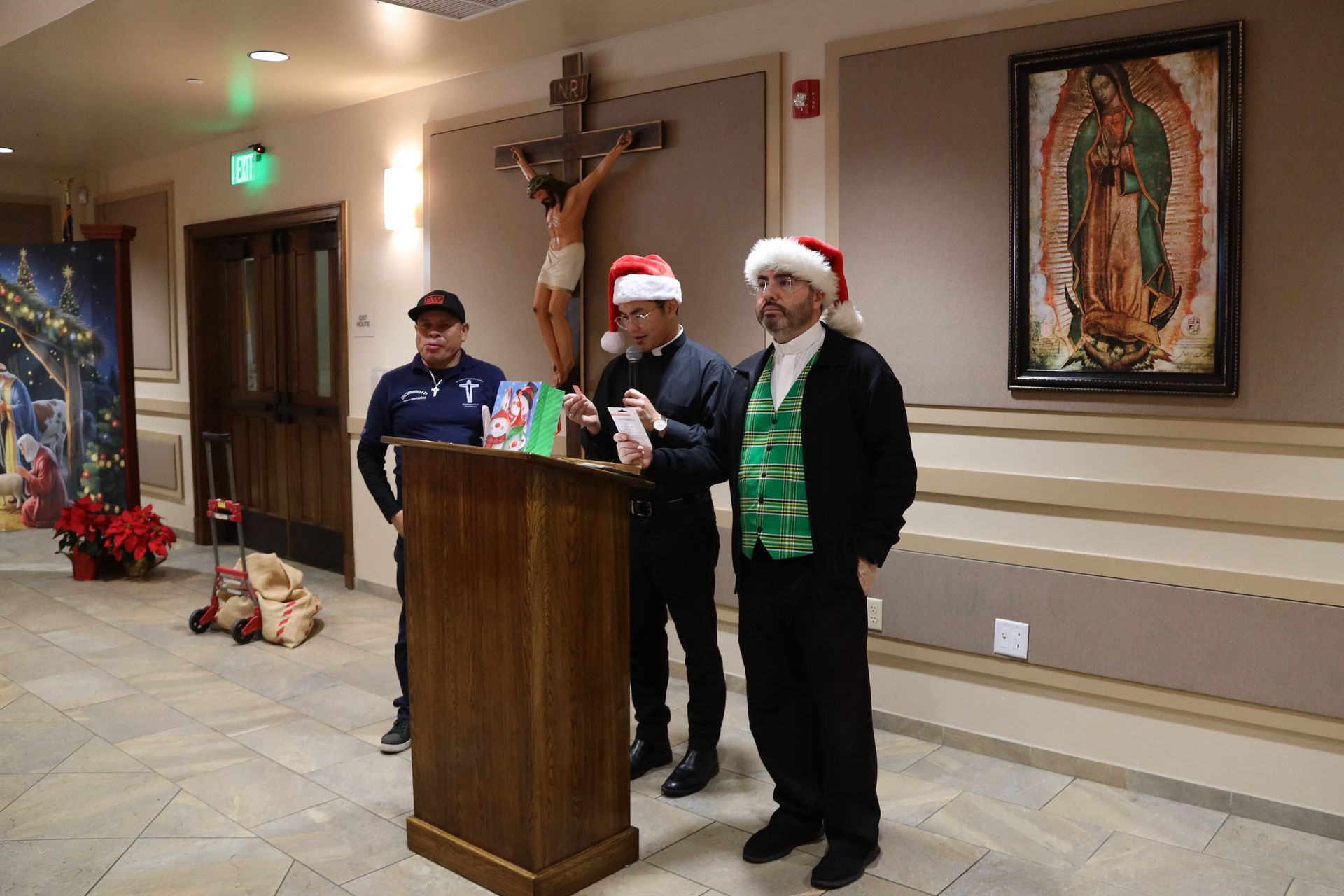 Three men in Santa hats at a podium in a hall, with a crucifix and Christmas decorations nearby