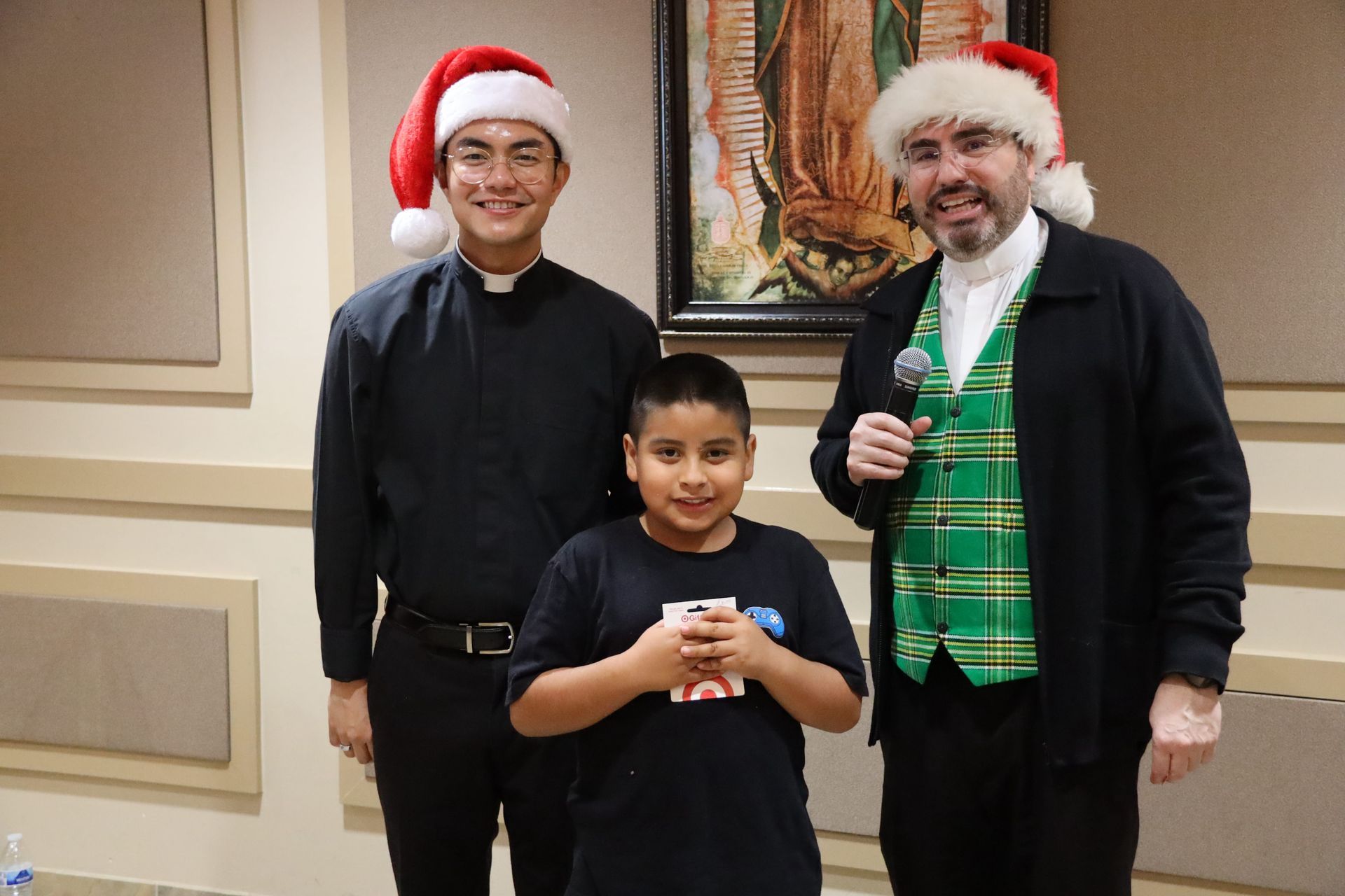 Three people in Santa hats posing indoors, one holding a microphone and one holding a small cup.
