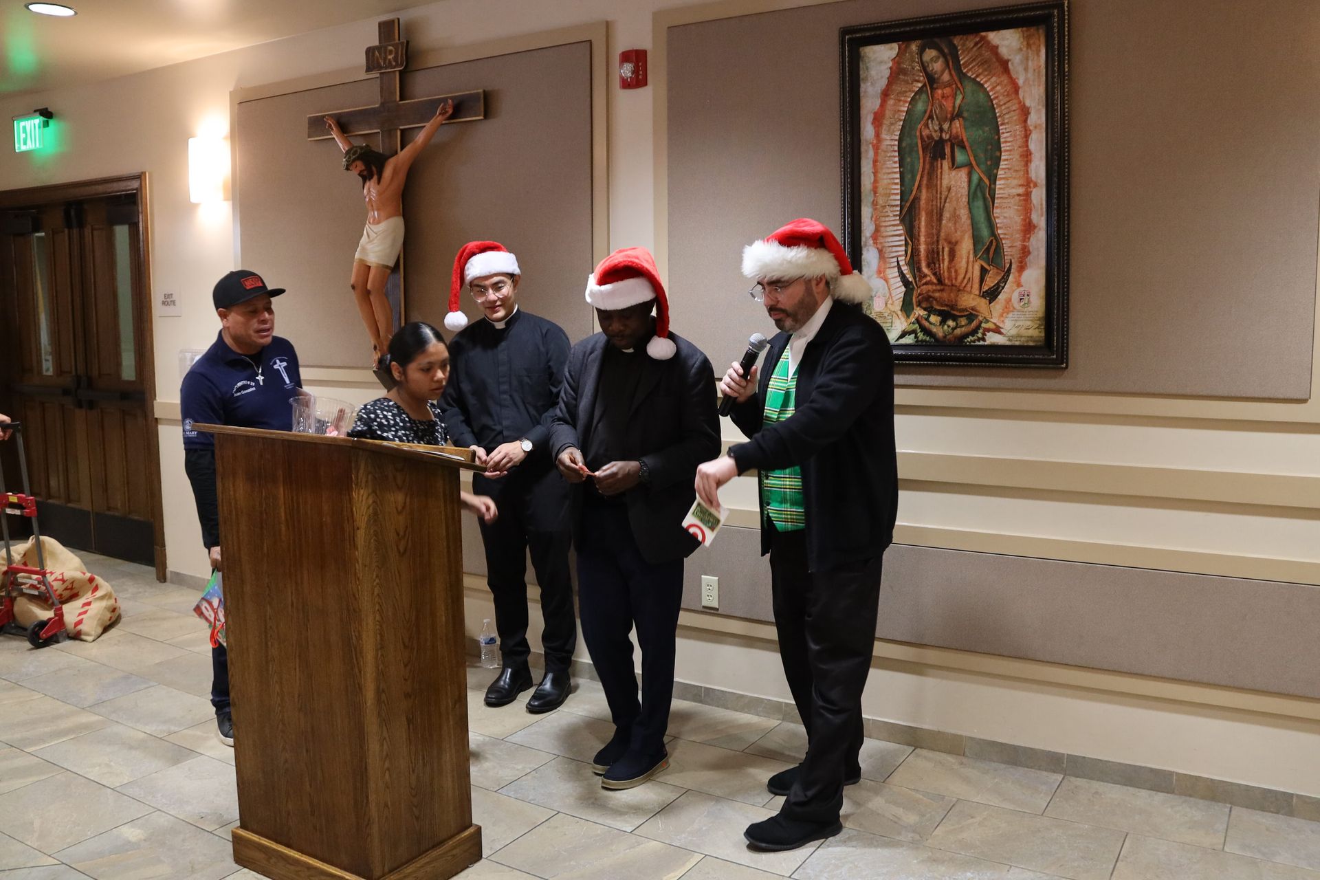 People in Santa hats stand at a podium beside a crucifix and religious artwork in a hall.