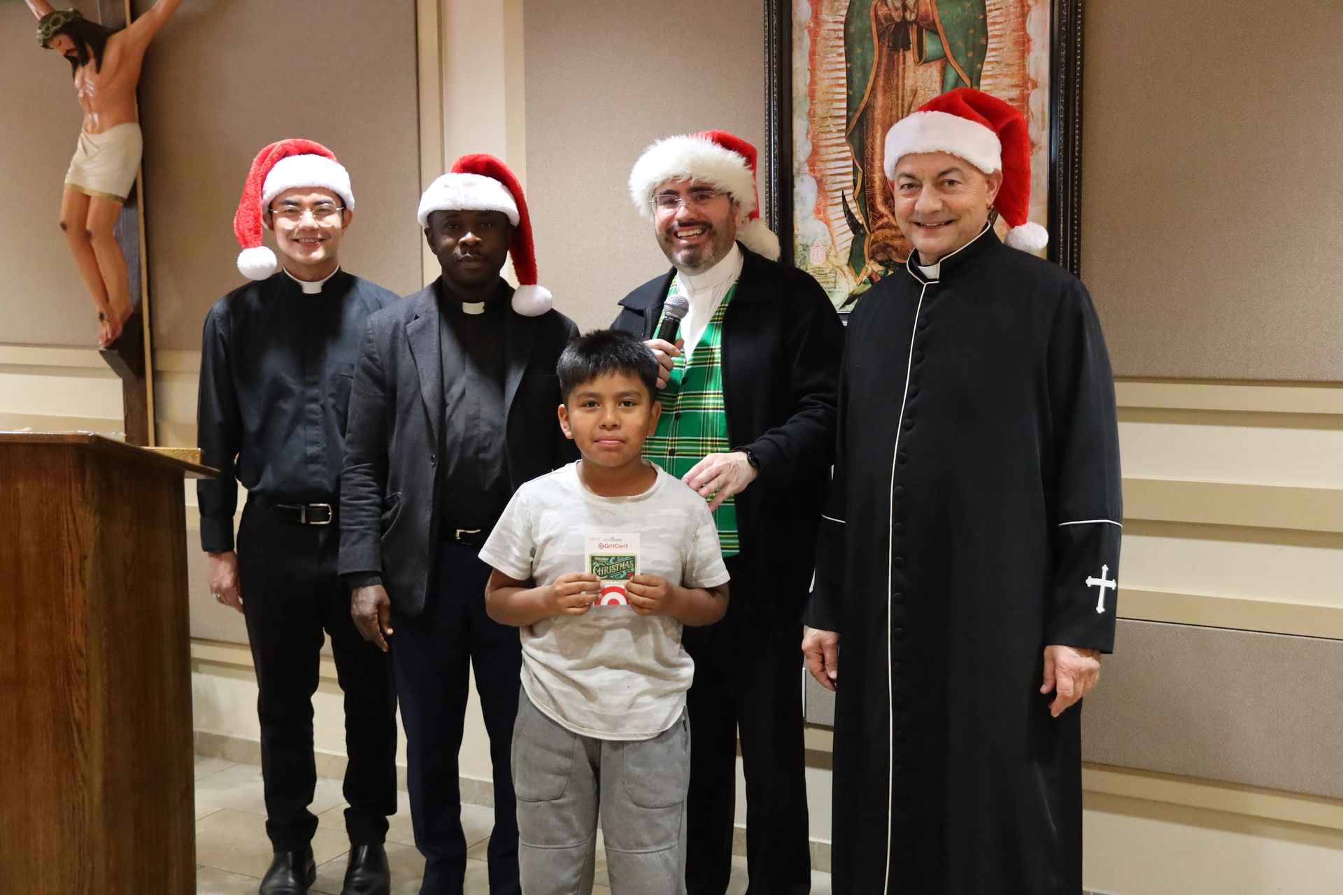 Four people in Santa hats stand behind a boy holding a small item indoors.