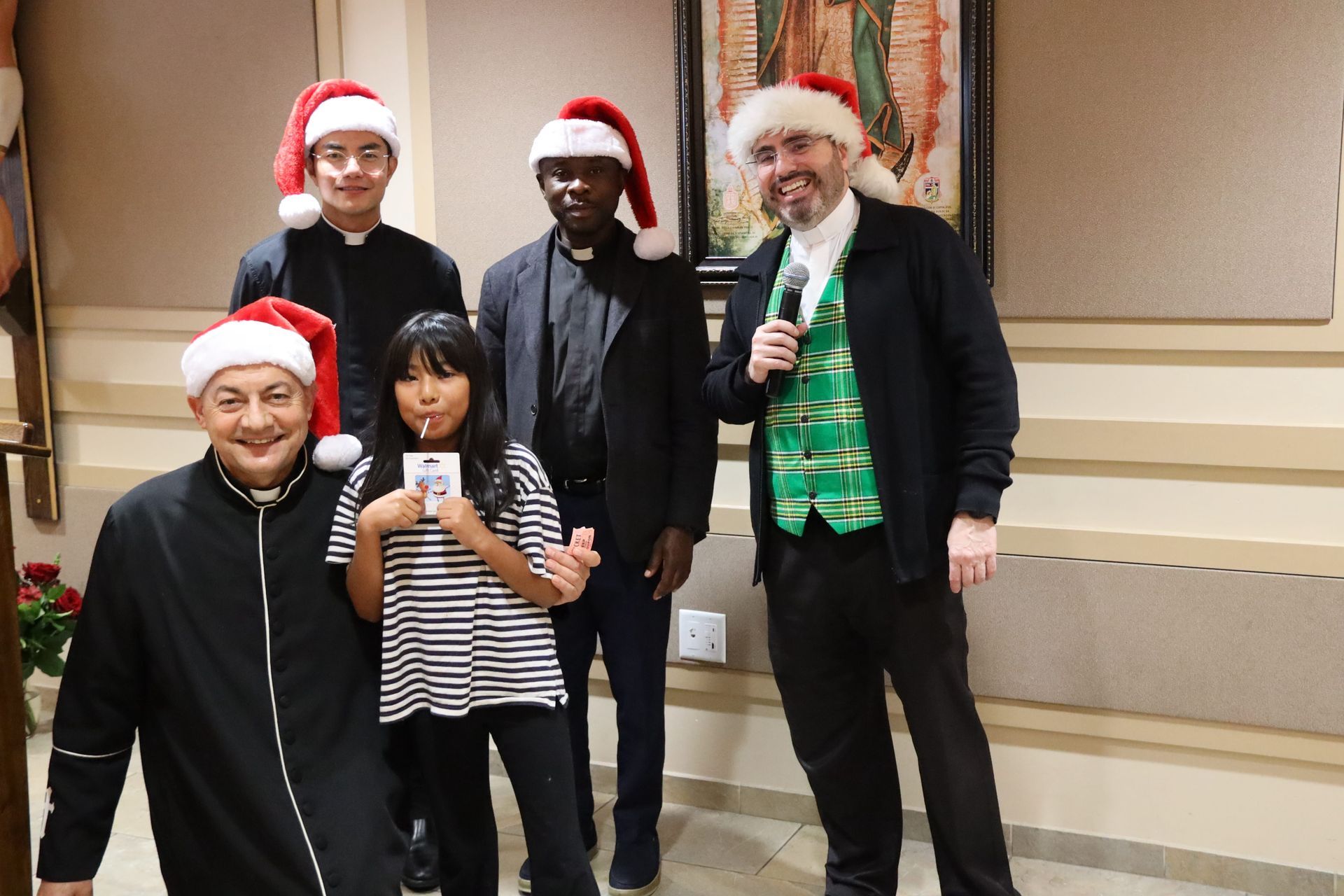 Five people in Santa hats posing indoors, one holding a microphone and a child holding a phone