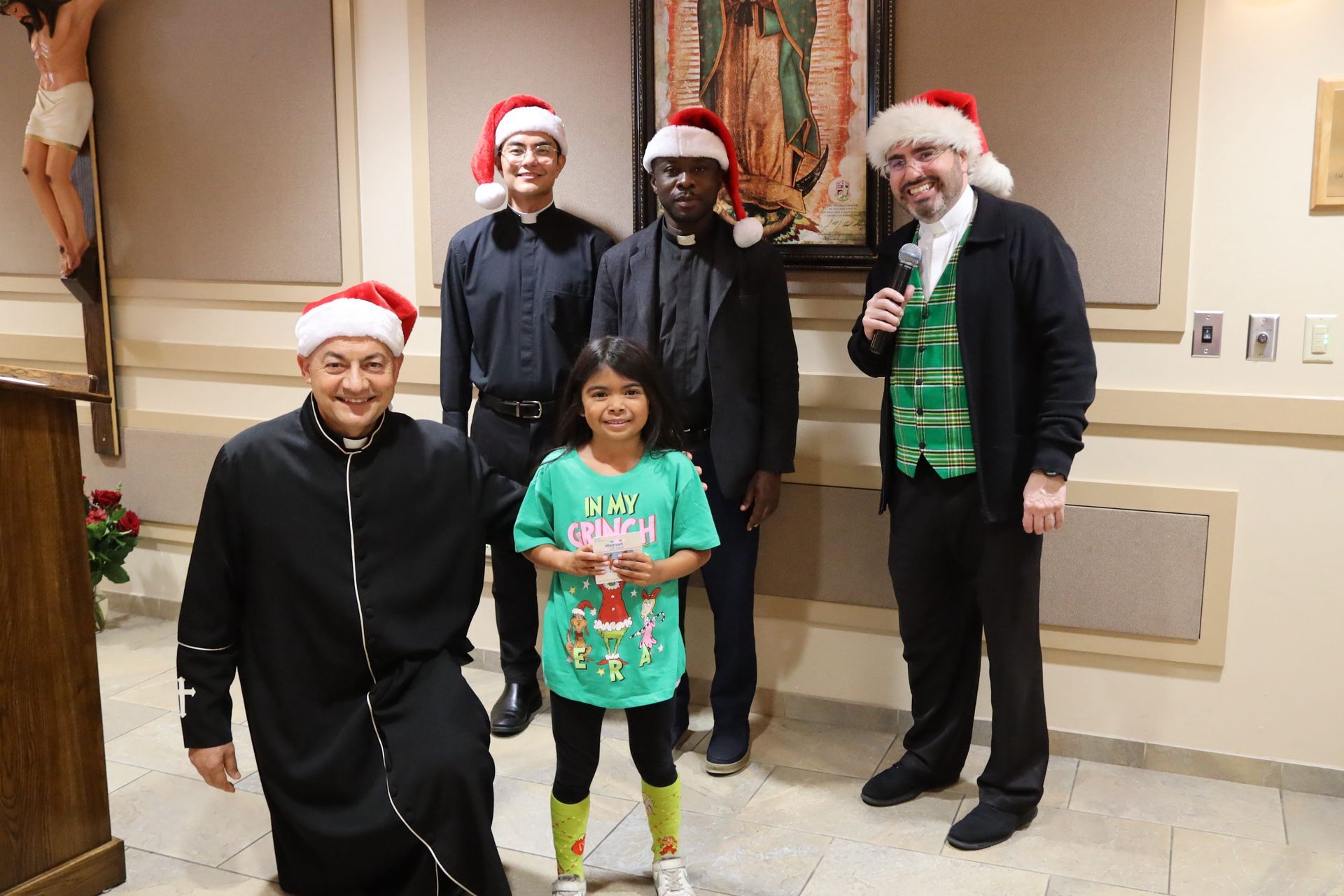 Five people in Santa hats posing indoors, one holding a microphone and one child holding a gift.