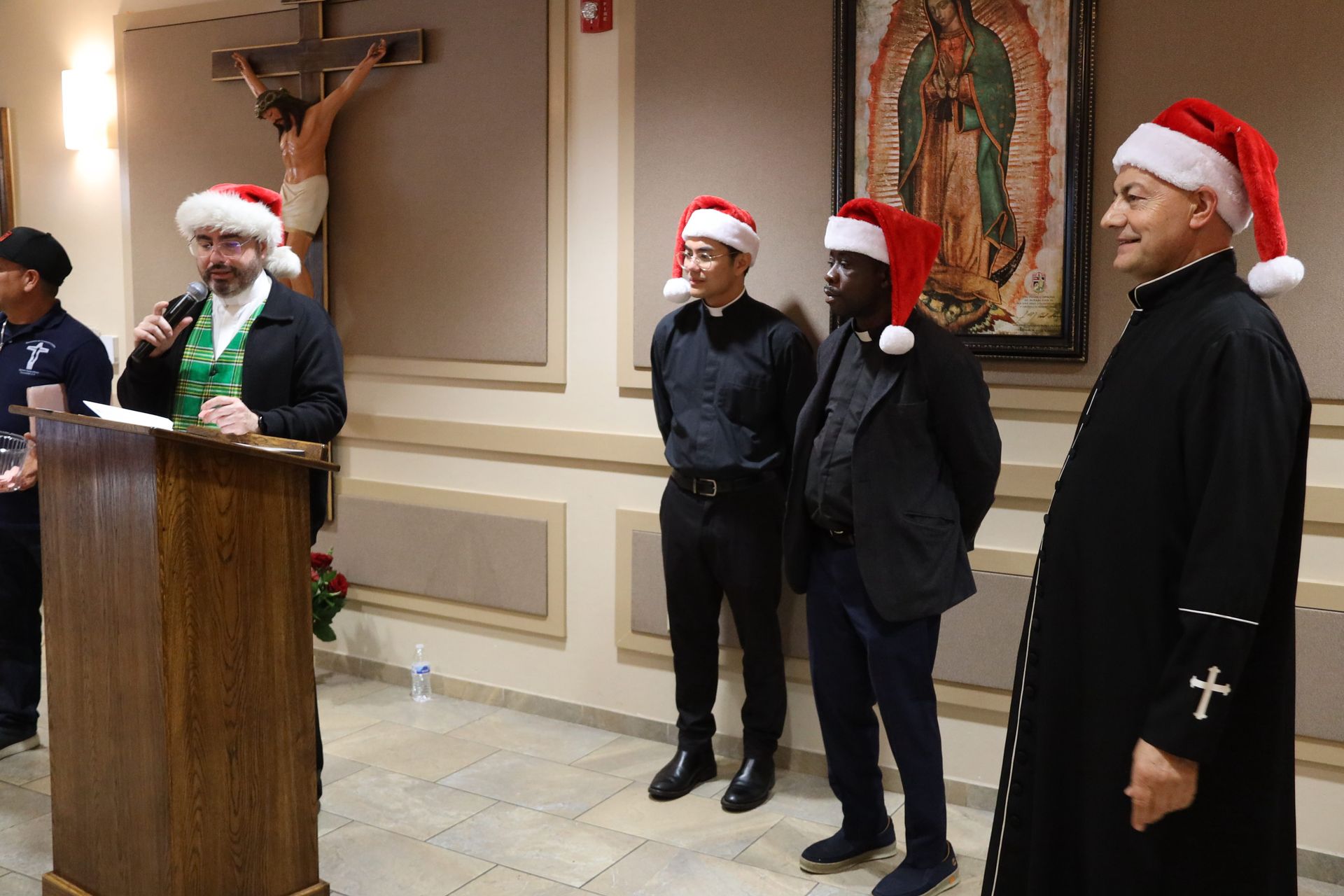 People in Santa hats standing at a podium in a church hall with a crucifix on the wall