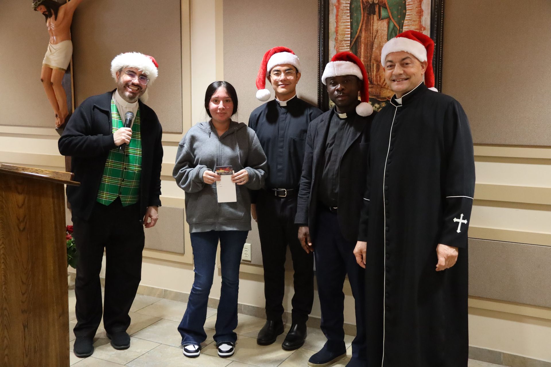 Five people posing indoors, four wearing Santa hats; one holds a cup and another wears a long black coat.