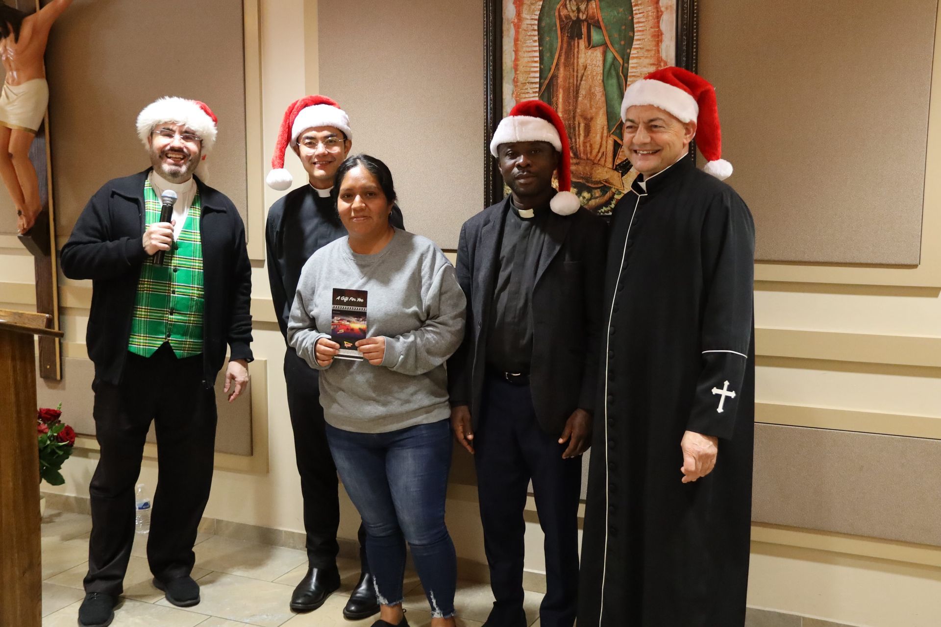 Five people in Santa hats pose indoors, holding drinks and smiling near a wall.