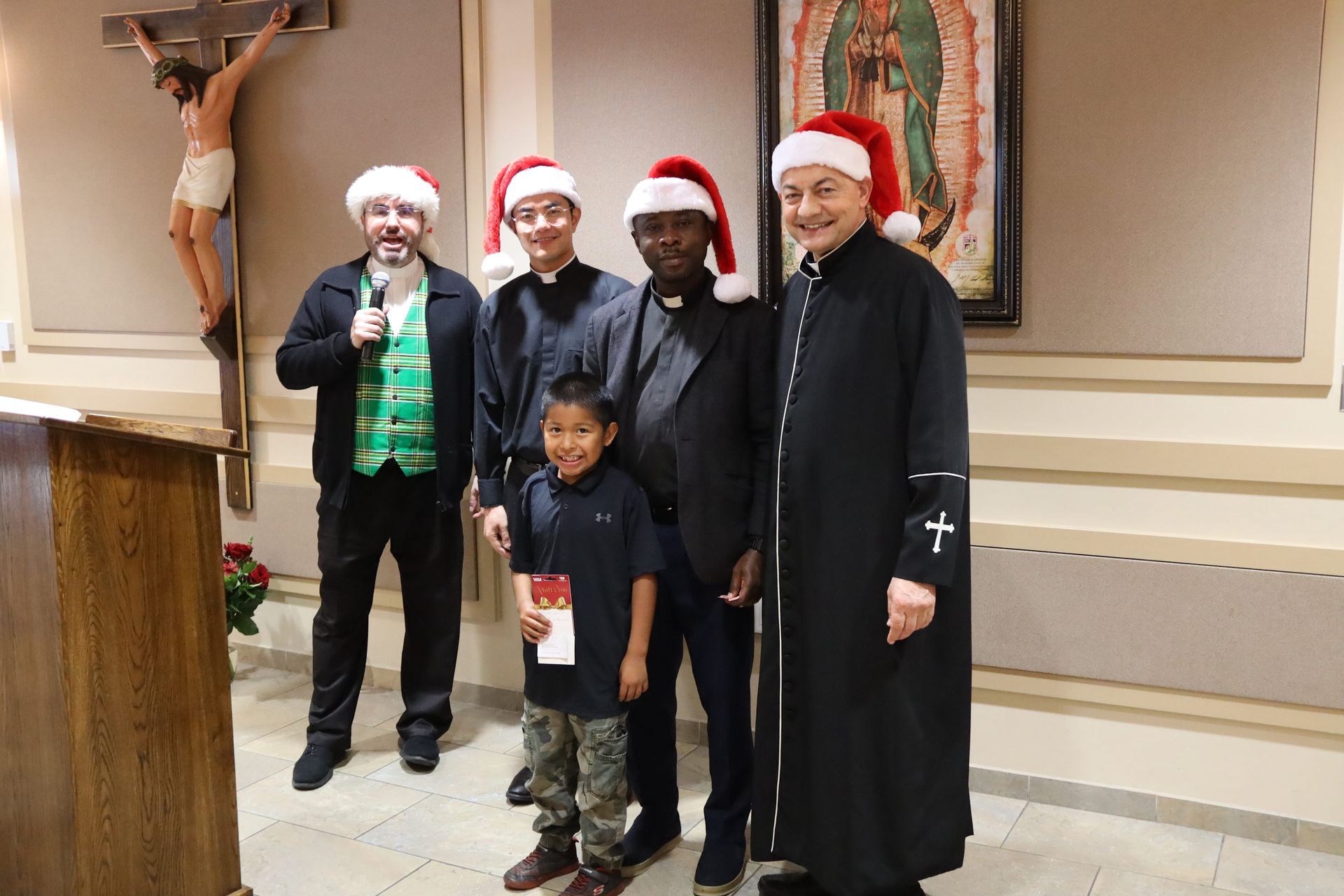 Five people in Santa hats posing indoors, with a crucifix on the wall and a child in front