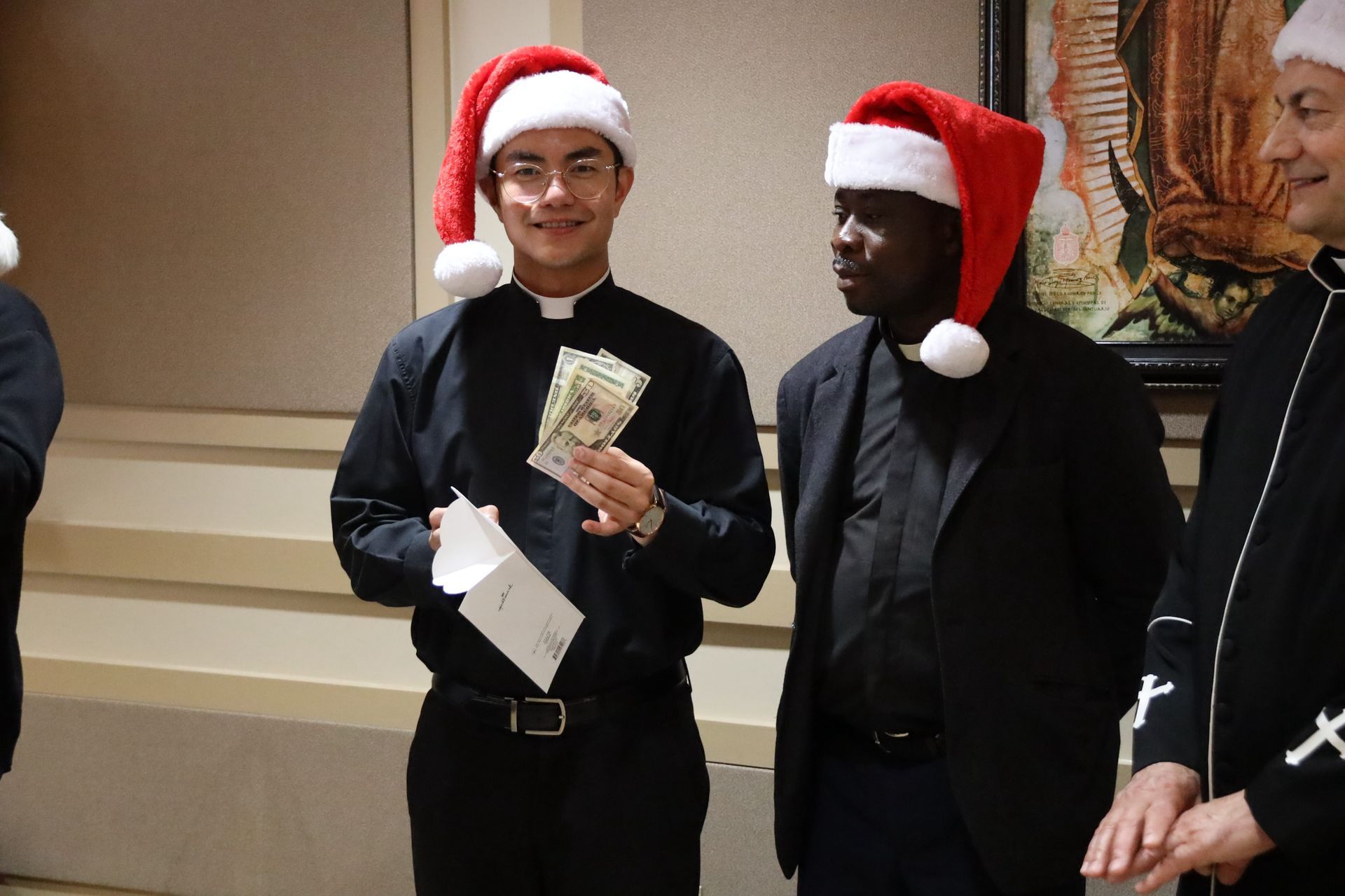 Three people in Santa hats stand indoors, one holding cash and an envelope.