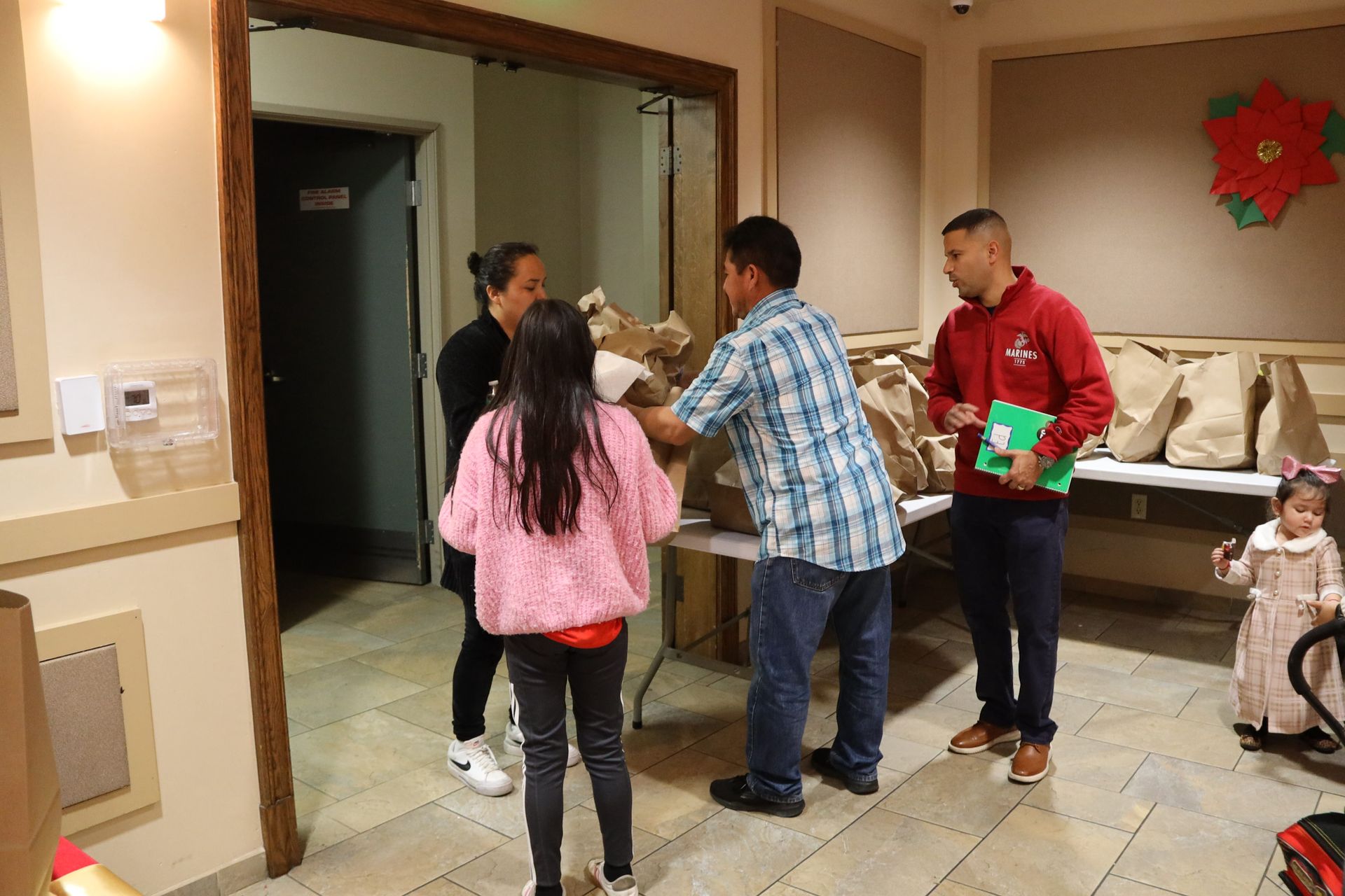 People exchanging gifts in a hallway, with a child nearby and wrapped presents on the floor.