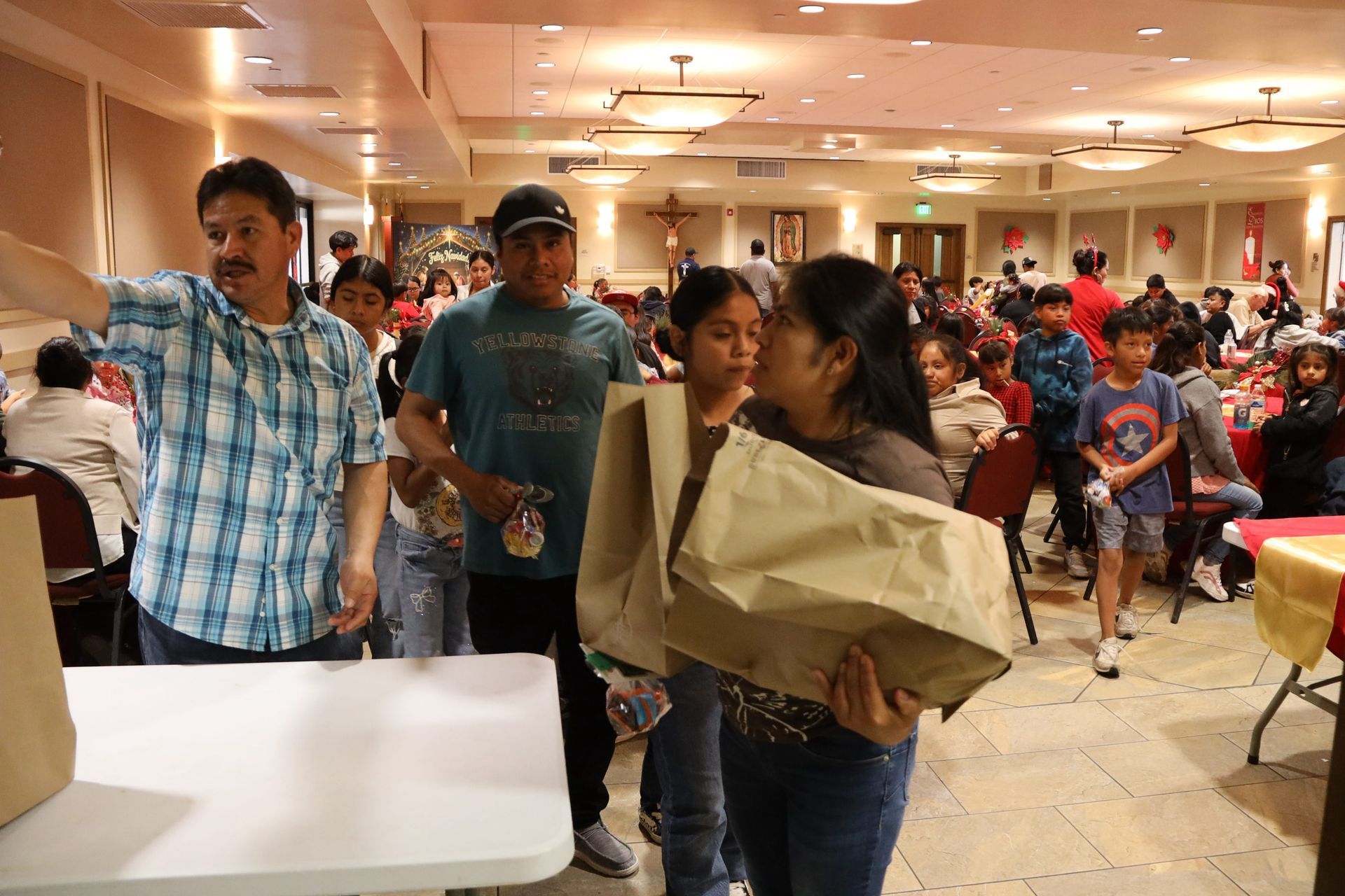 People carrying boxes in a crowded indoor hall with tables and chairs spread around.