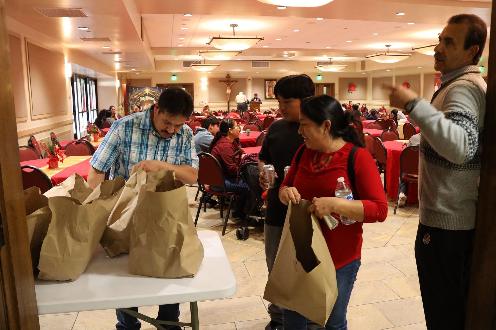 People packing brown paper bags at a crowded banquet hall table with red chairs and warm lighting