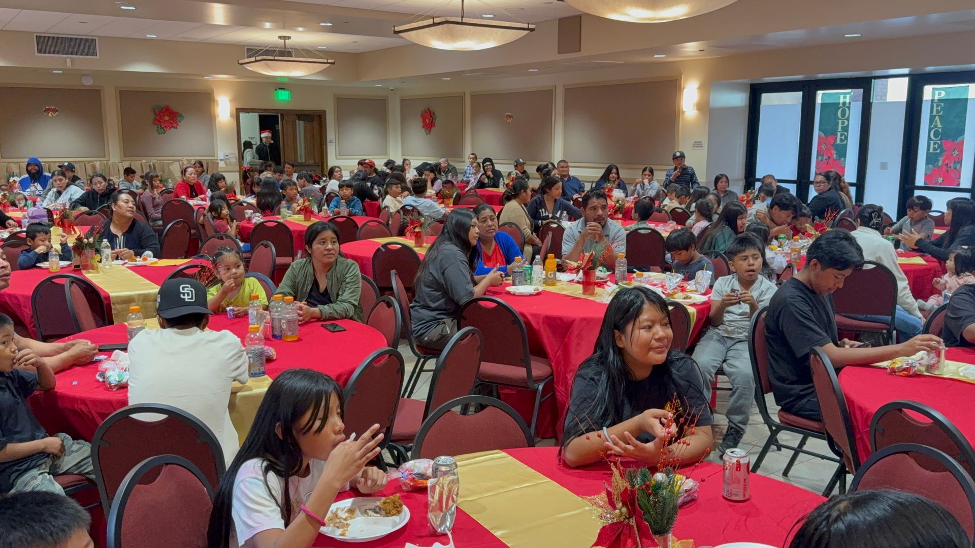 Crowded banquet hall with people seated at red-covered tables eating a meal.
