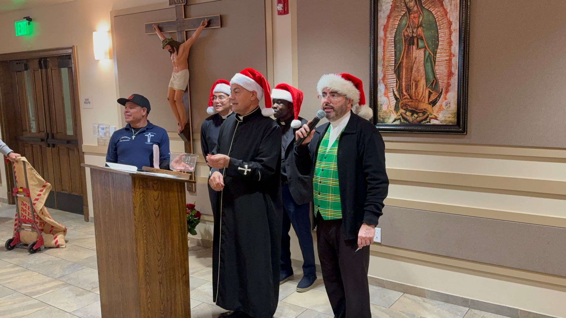 Three men in Santa hats stand at a church podium, one speaking into a microphone.