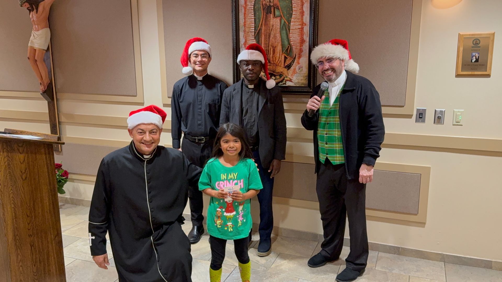 Five people in Santa hats pose indoors, with a child in a green holiday shirt in front.