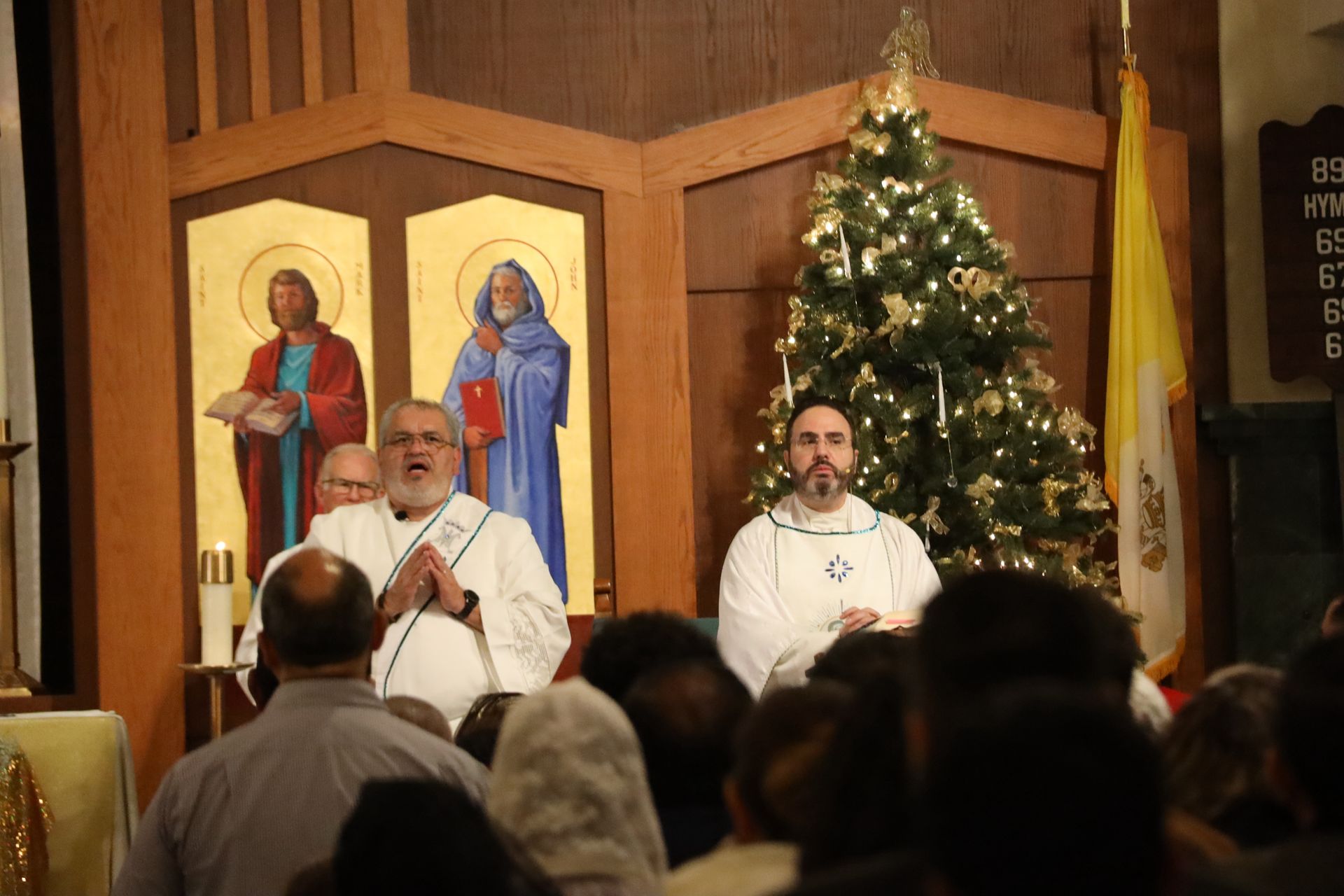 Church service with two clergy at the altar, icons behind them, and a lit Christmas tree to the side
