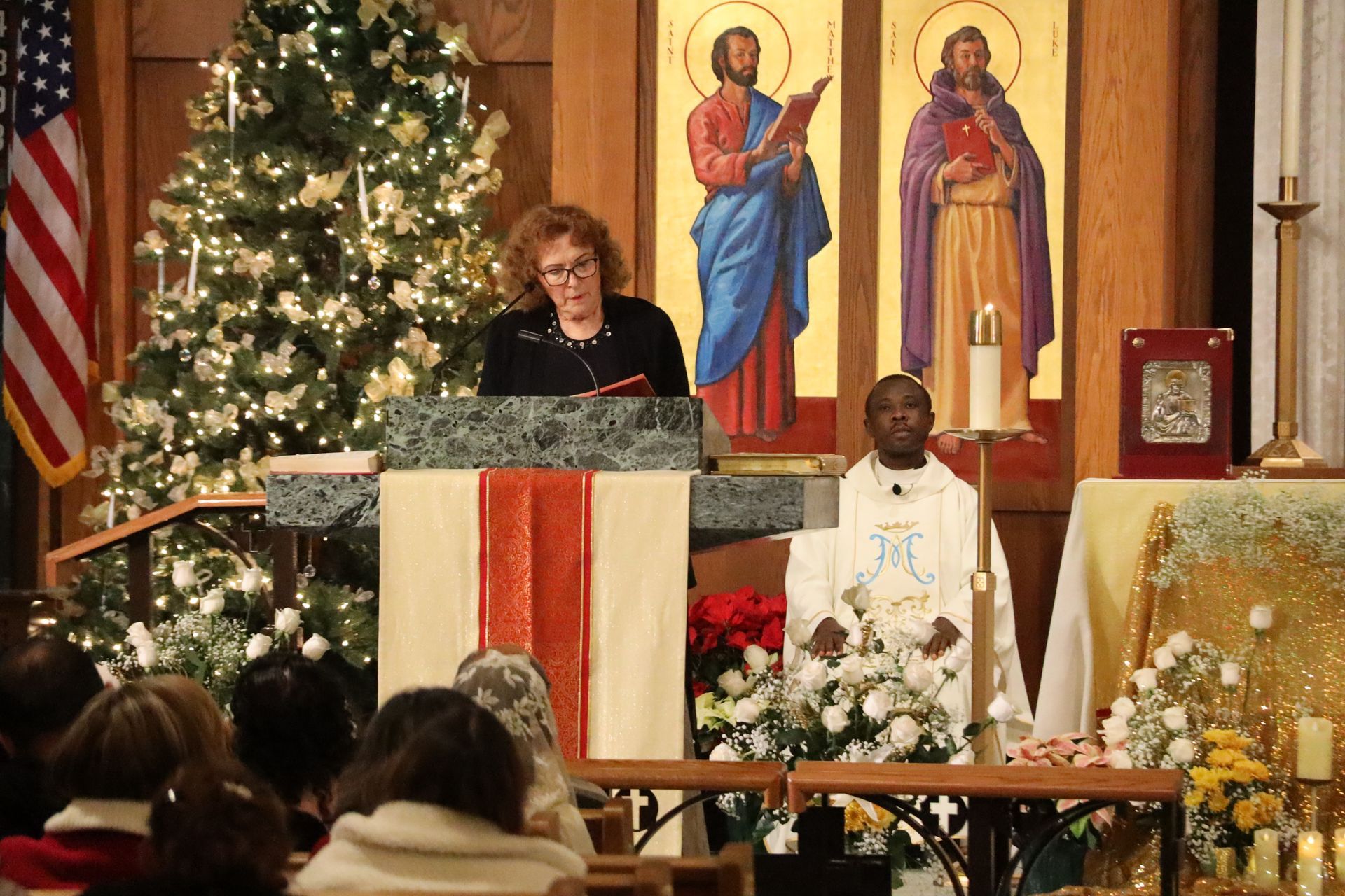 Woman speaking at church lectern beside lit Christmas tree and altar