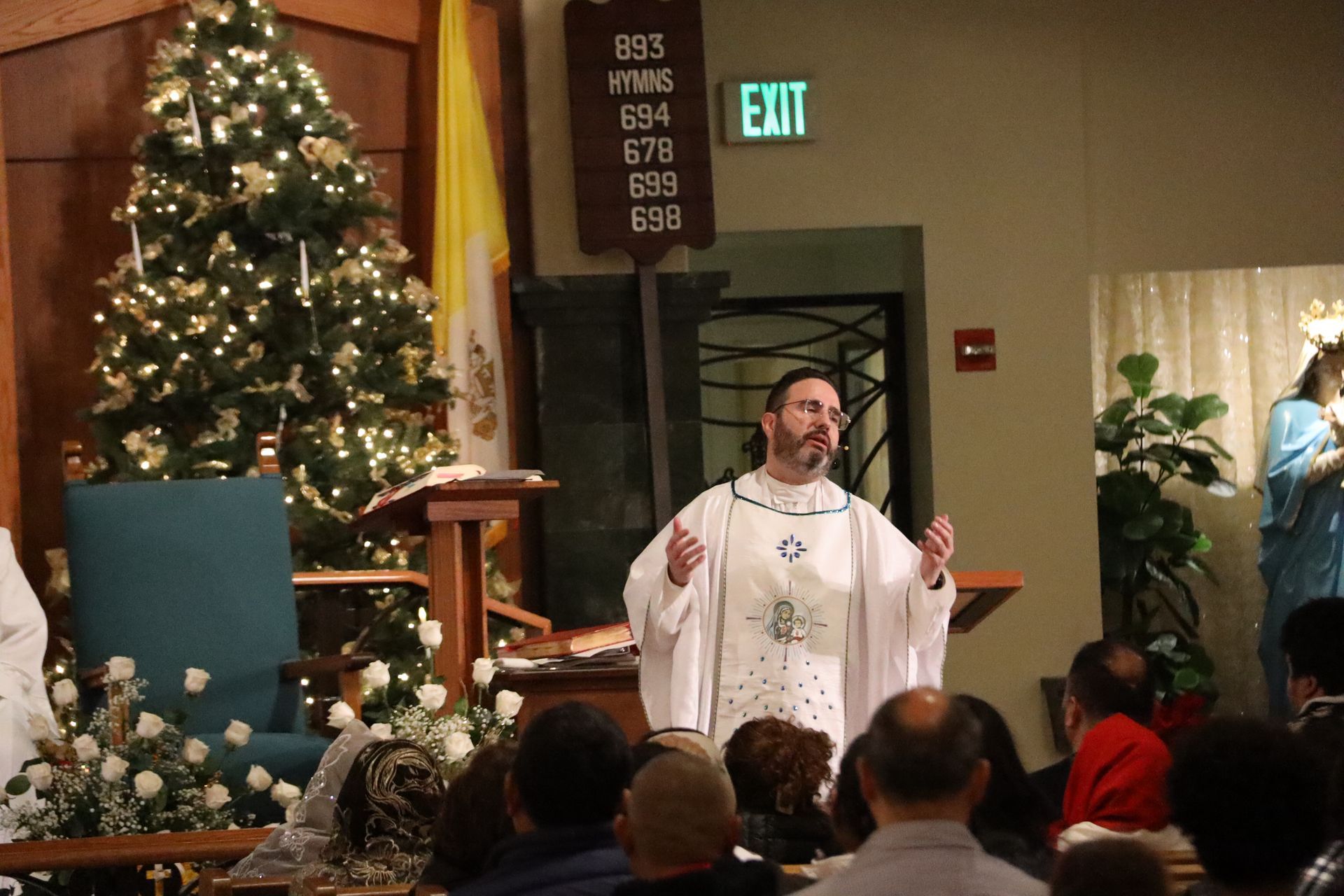 Priest in white robe speaking at the altar during a Christmas church service, with decorated tree nearby