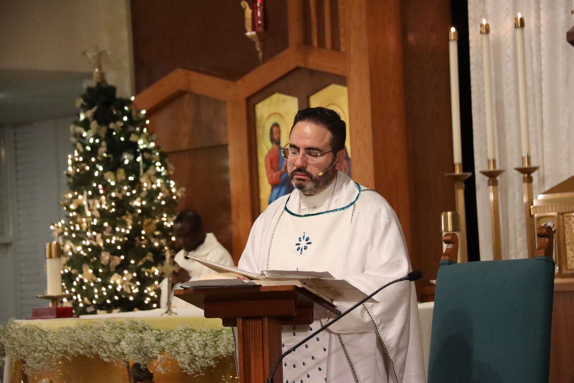 Clergyman in white vestments speaking at a lectern in a church beside a lit Christmas tree.