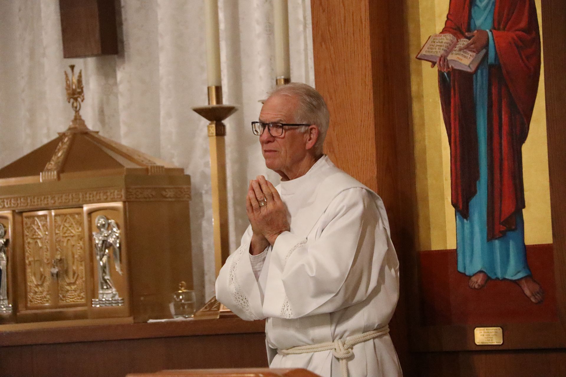 Priest in white vestments prays beside an altar in a church.