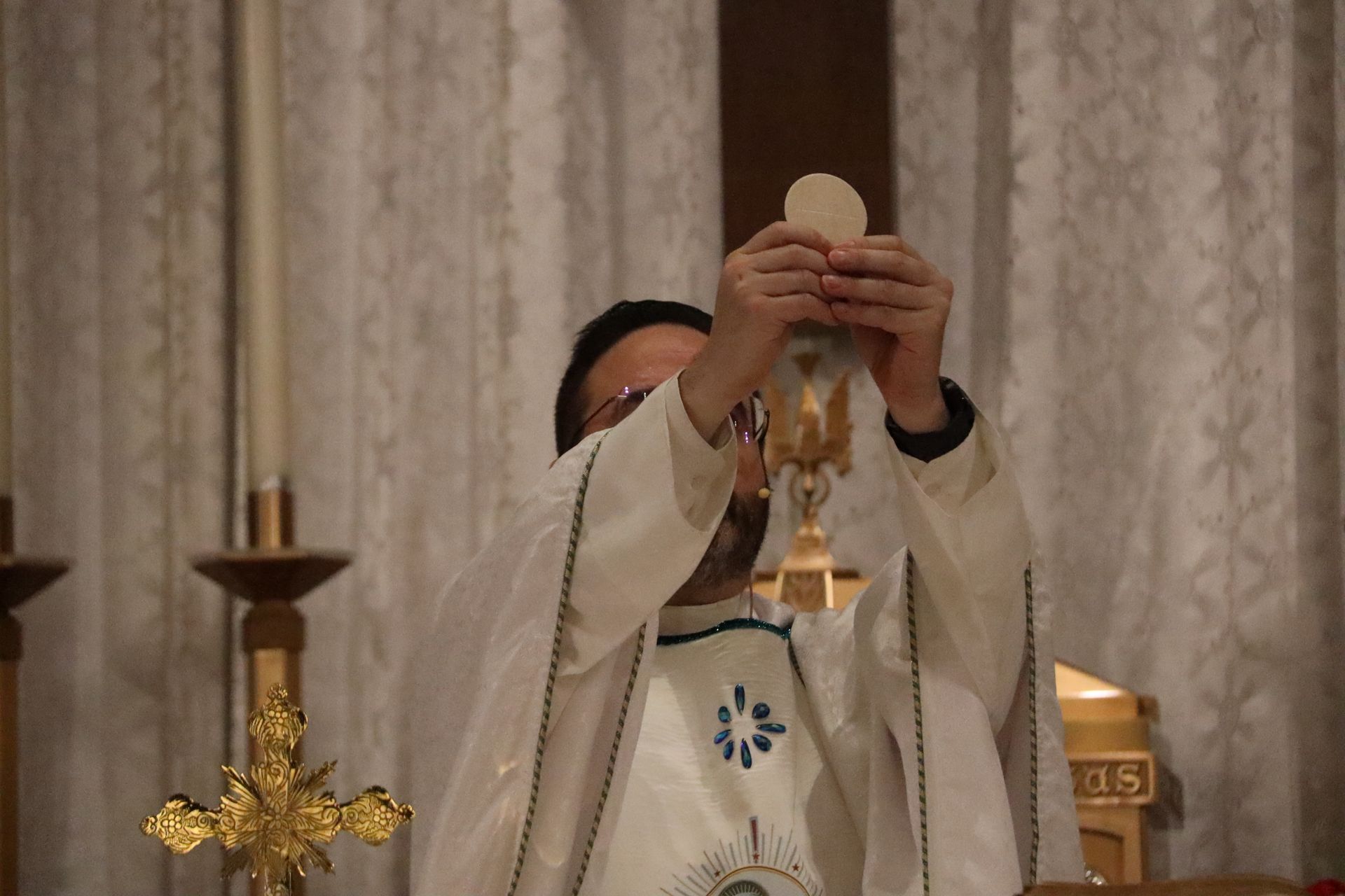 Priest in white robes elevates a communion wafer during Mass before altar candles