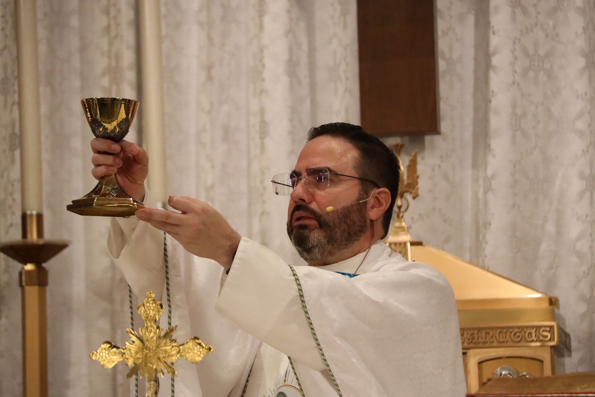 Priest in white vestments elevates a gold chalice during a church ceremony.