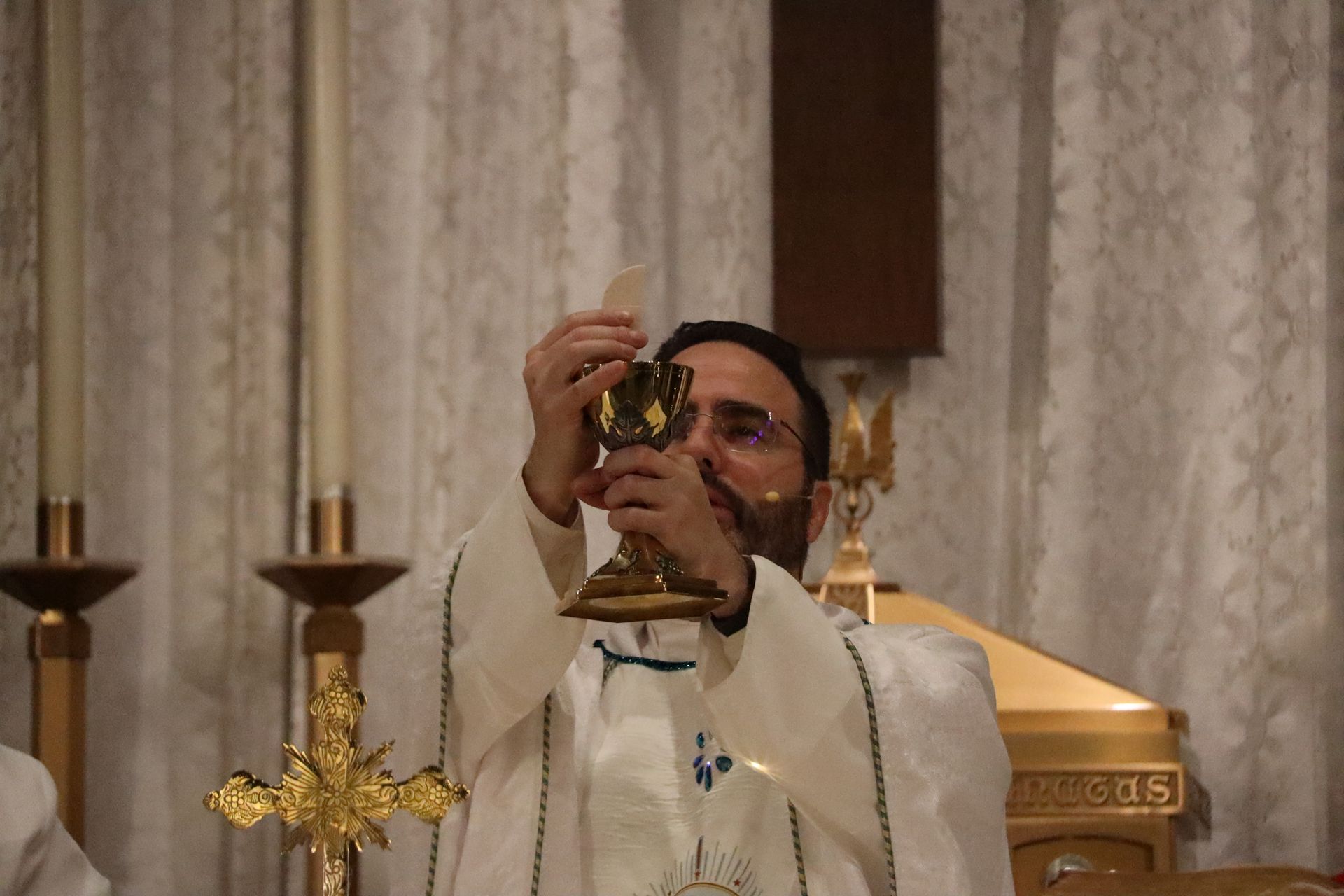 Priest elevates a chalice during Catholic Mass before an altar with candles and lace curtains