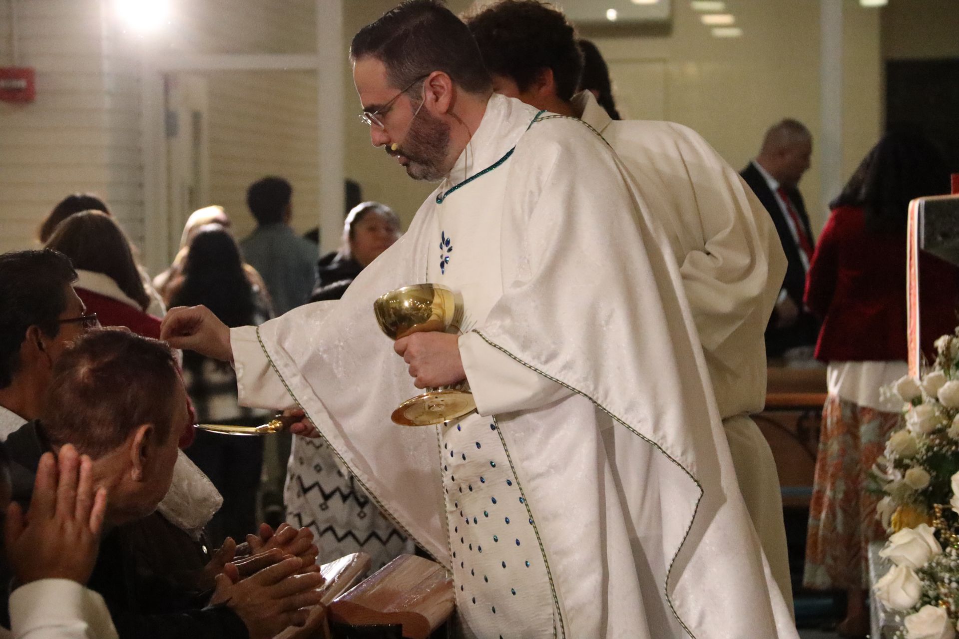 Priest in white vestments distributing communion to seated worshippers in a church