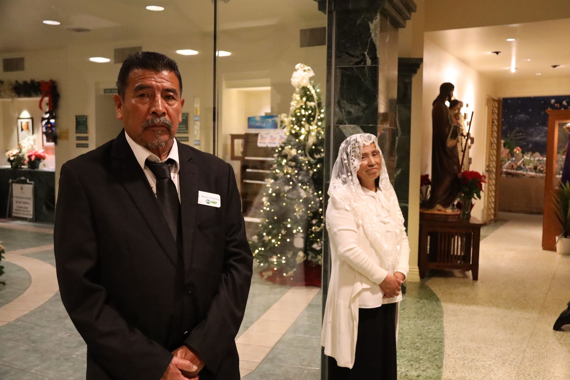 Two hotel employees in black suits stand in a decorated lobby beside a lit Christmas tree.