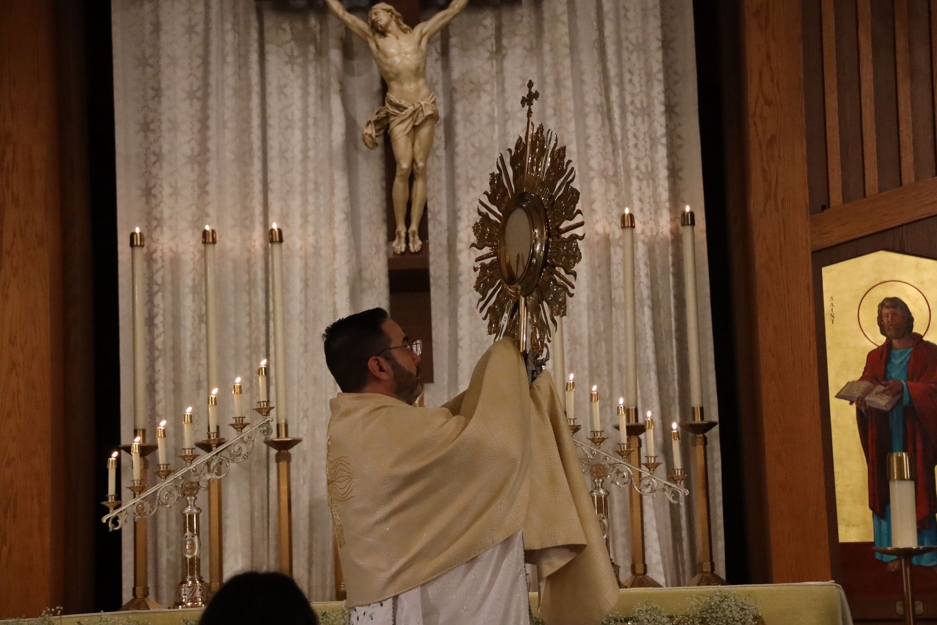 Priest elevates a golden monstrance before the altar beneath a crucifix in a church.