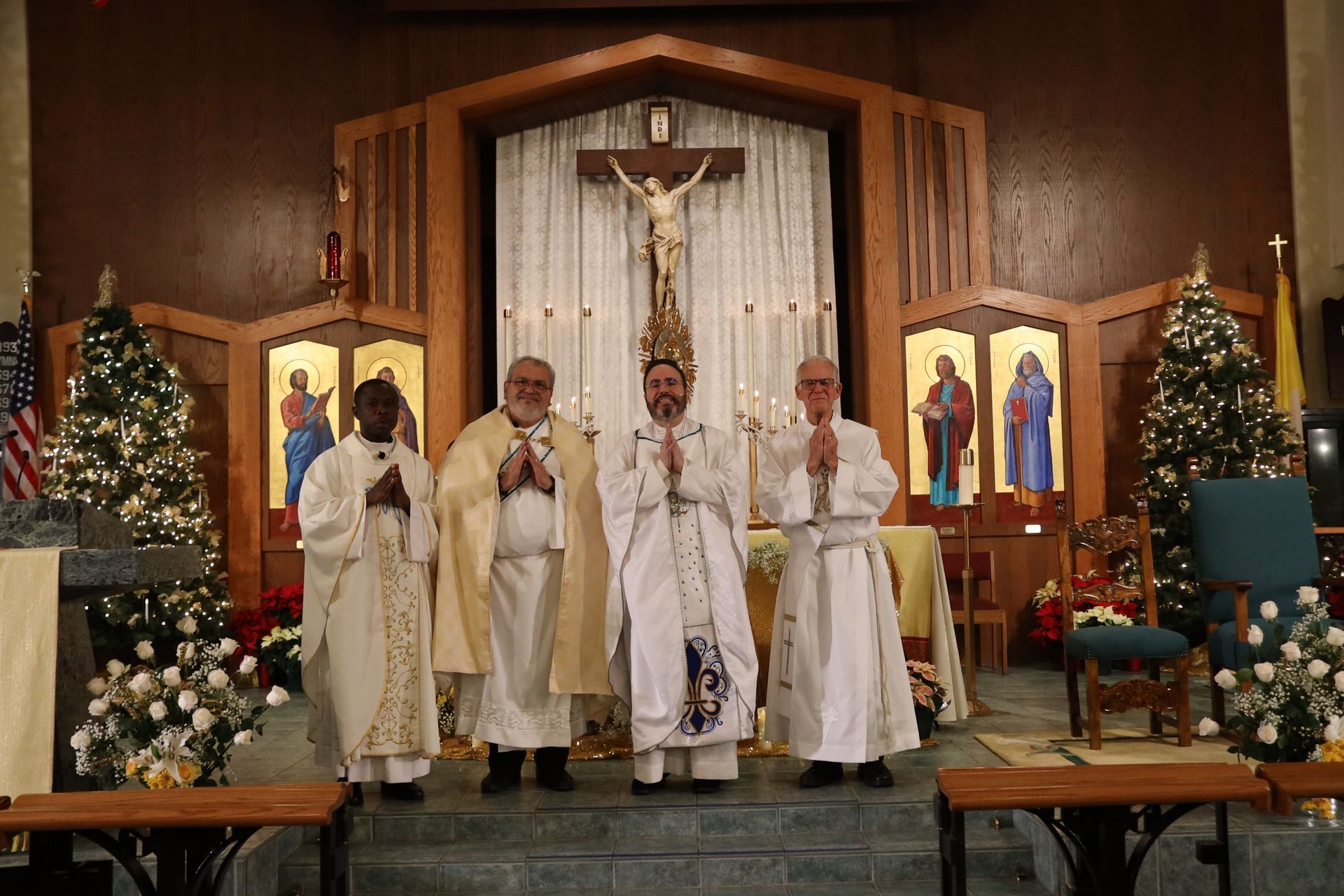 Four priests in white vestments stand at a church altar beneath a crucifix and Christmas decorations.