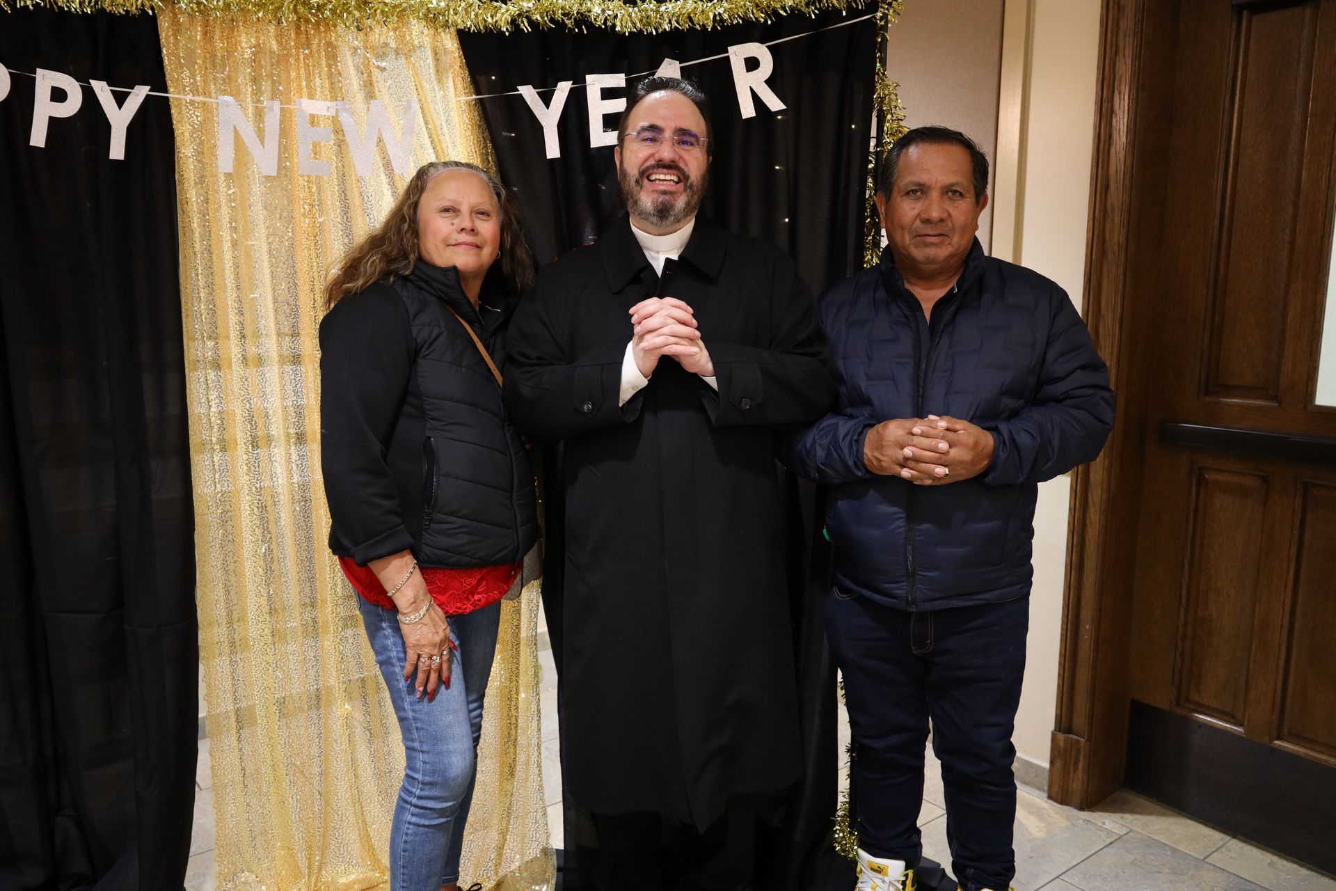 Three people posing indoors in front of a “Happy New Year” backdrop, smiling at the camera.