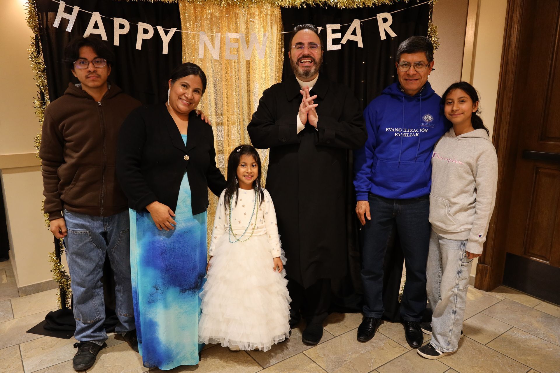 Family standing indoors in front of a “Happy New Year” banner, with two children and festive decor.