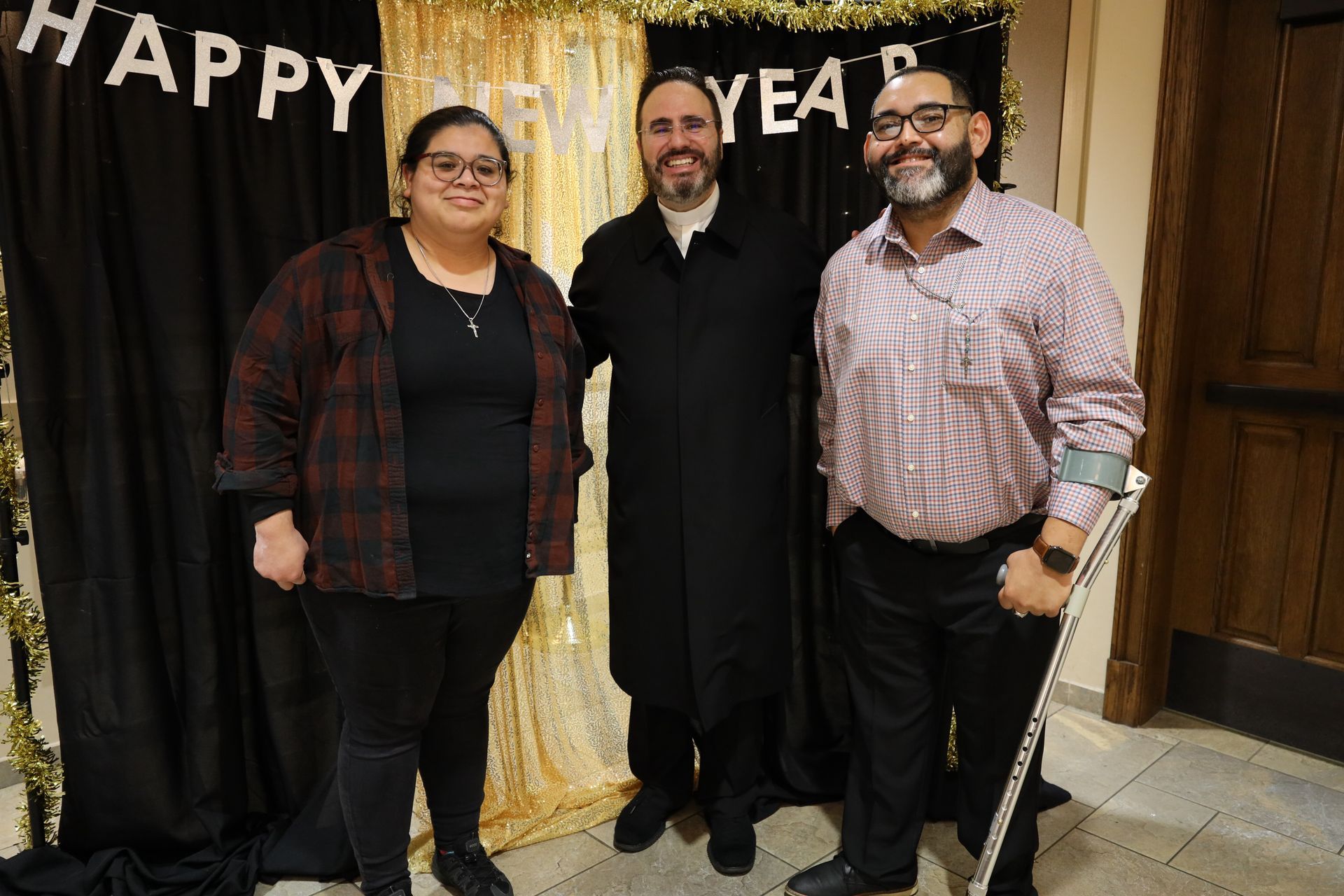 Three people posing at a celebration backdrop with gold curtains and “HAPPY NEW YEAR” banner