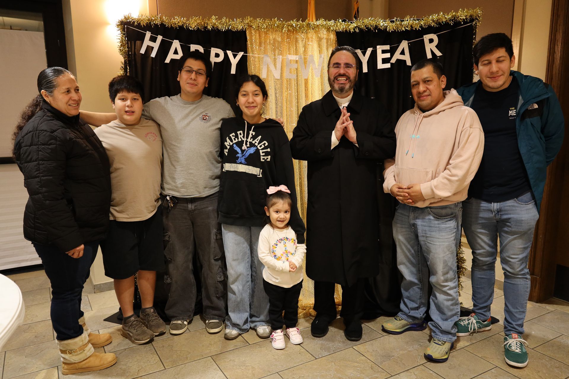 Group of people posing indoors in front of a gold “HAPPY NEW YEAR” banner, smiling and standing together.