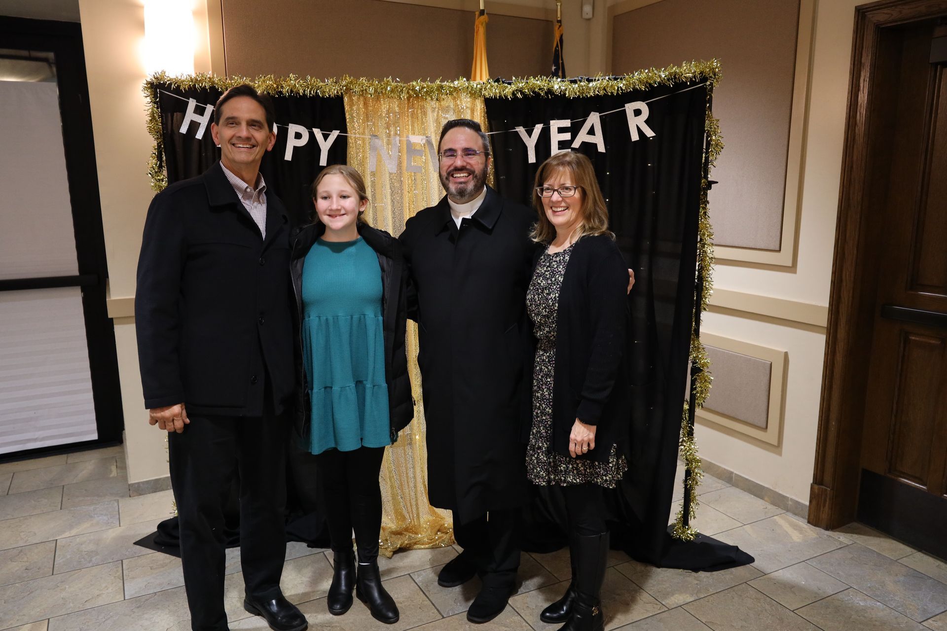 Four people posing at a New Year photo backdrop with gold tinsel and a black “Happy New Year” banner.