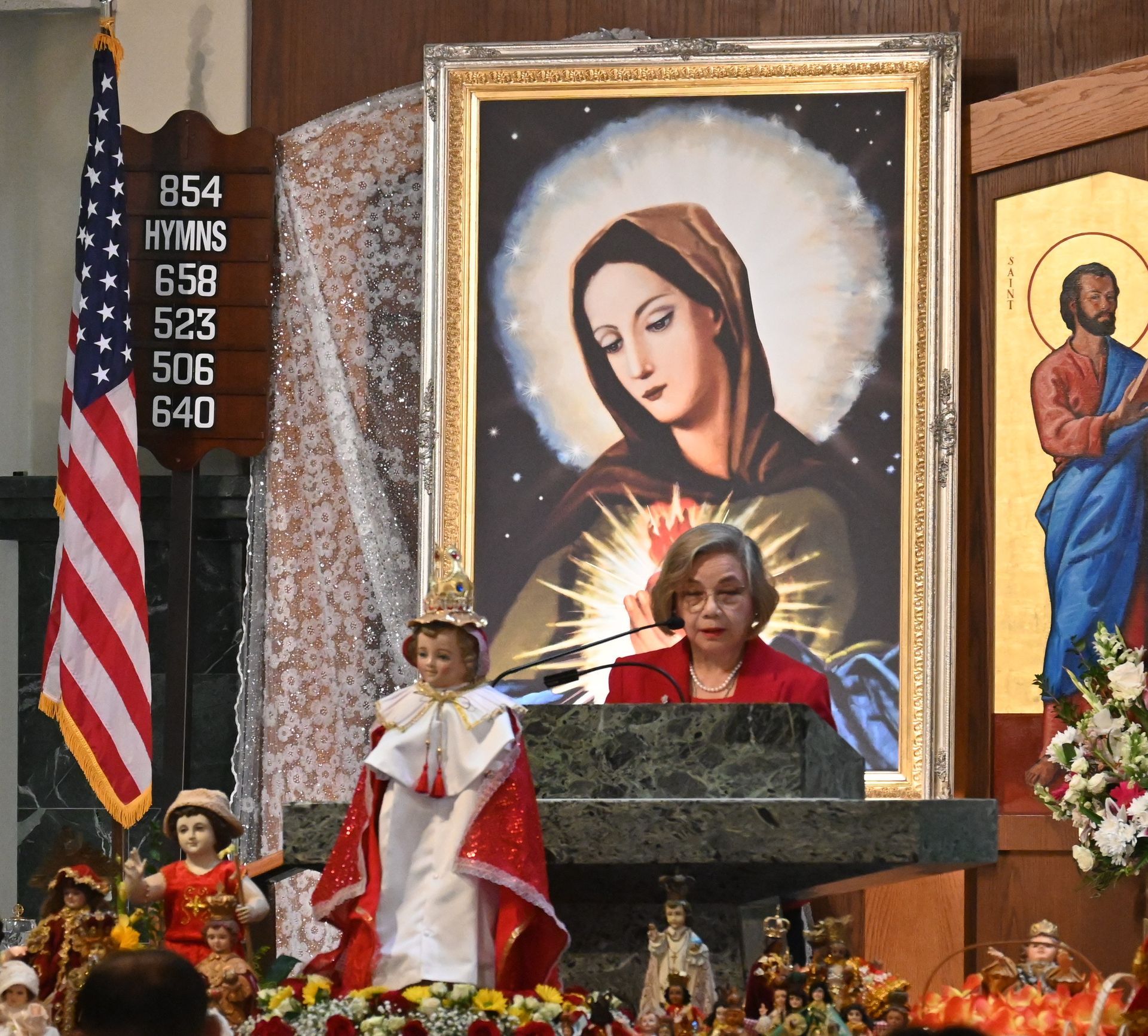 Woman speaking at a podium in a church, with a large framed image of the Virgin Mary behind her.