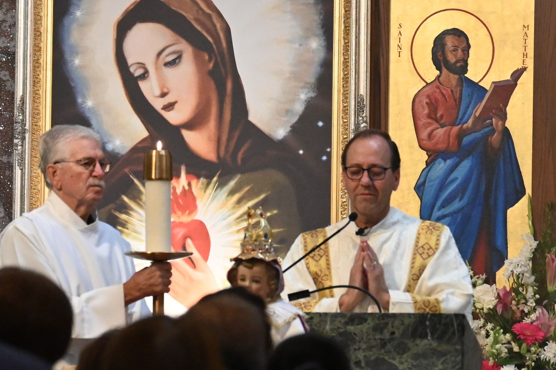 Two priests at a church altar before a Virgin Mary mural and a saint icon