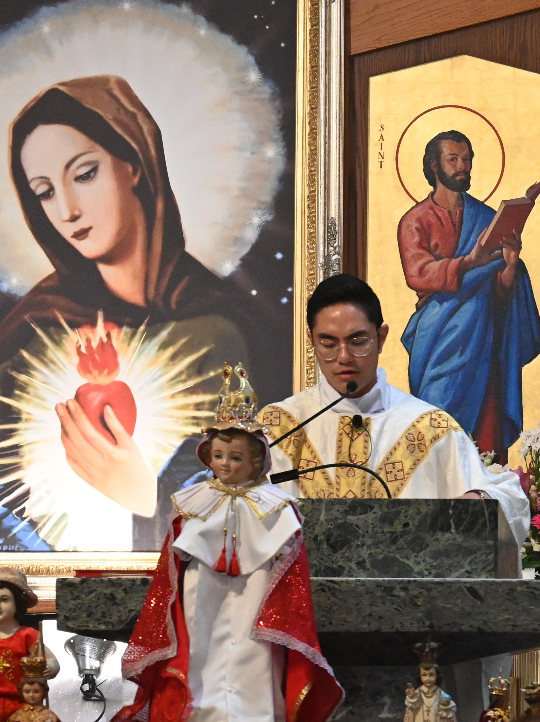 Man speaking at church lectern beside Marian and saint icons, with a small statue in front