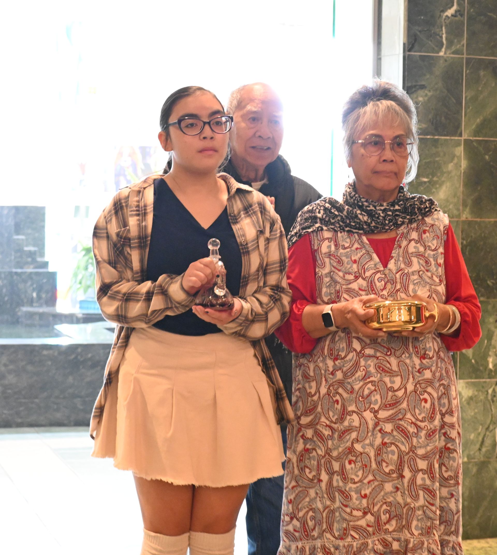 Three people standing indoors, two holding small gold objects, in front of a bright window.