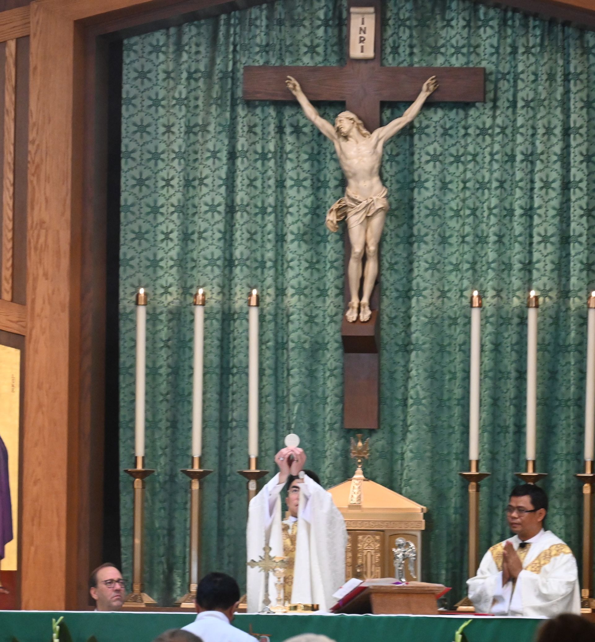 Priest at altar beneath large crucifix, with candles and green backdrop in a church sanctuary