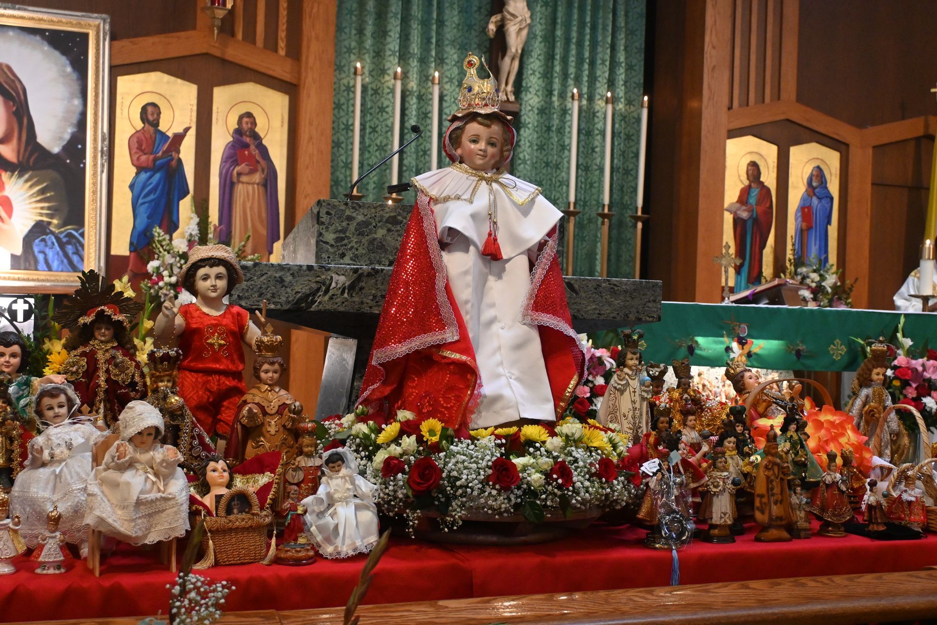 Religious altar display with a crowned child statue in white and red surrounded by flowers and smaller figurines