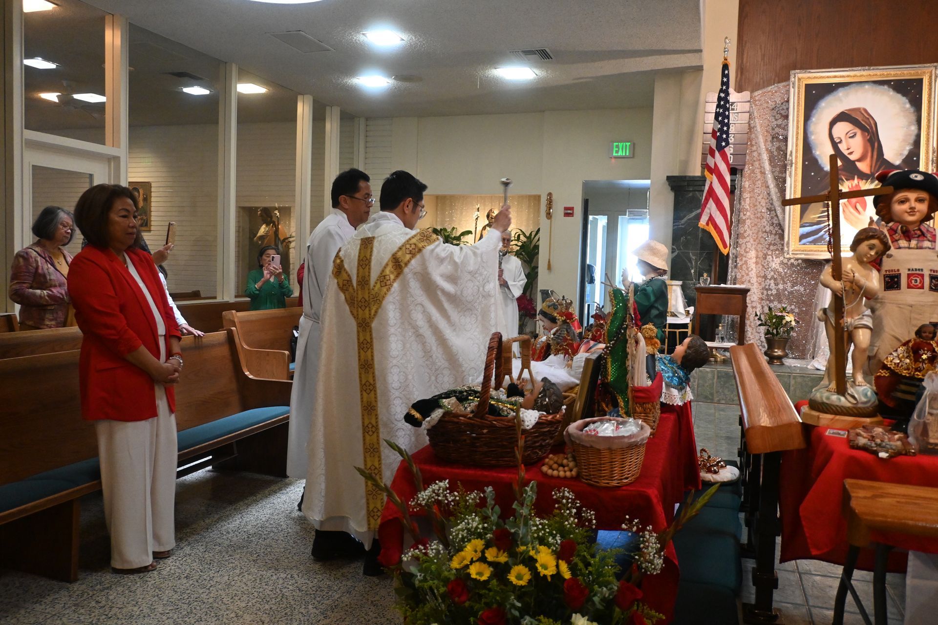 Priest in white robes conducts a church ceremony beside a decorated altar with flowers and candles.