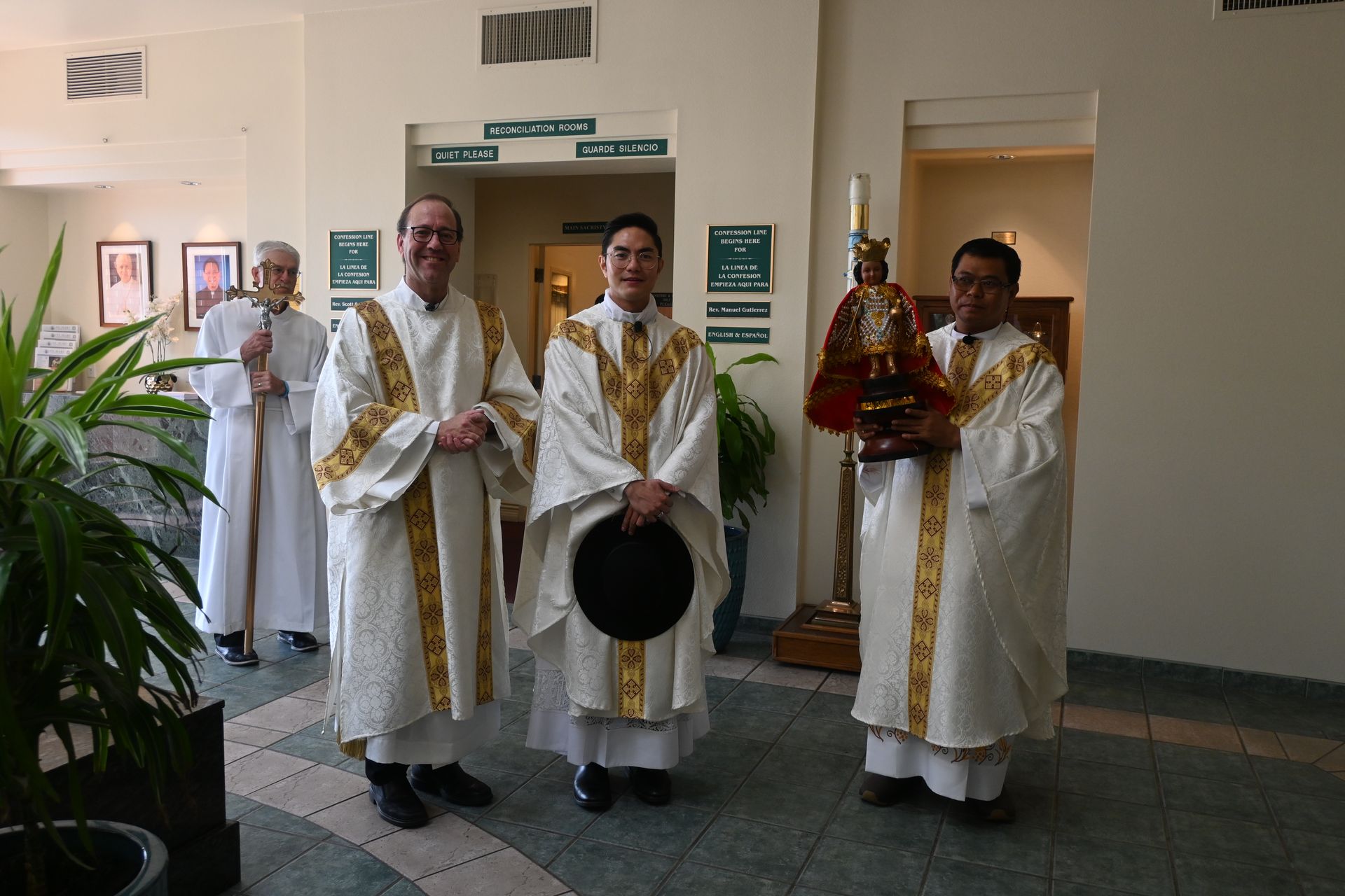 Three robed clergy stand indoors, one holding a red statue, in a hallway with wall art.