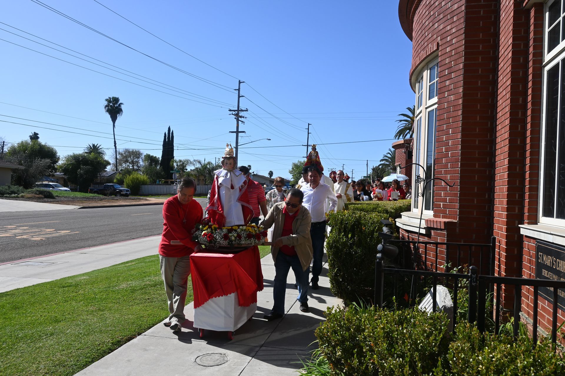 People gathered around a red-and-white table outside a brick building on a sunny sidewalk