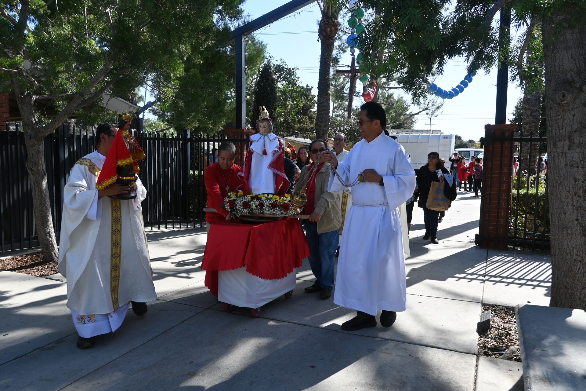 People in white robes carry a decorated red platform through a gated entrance outdoors.