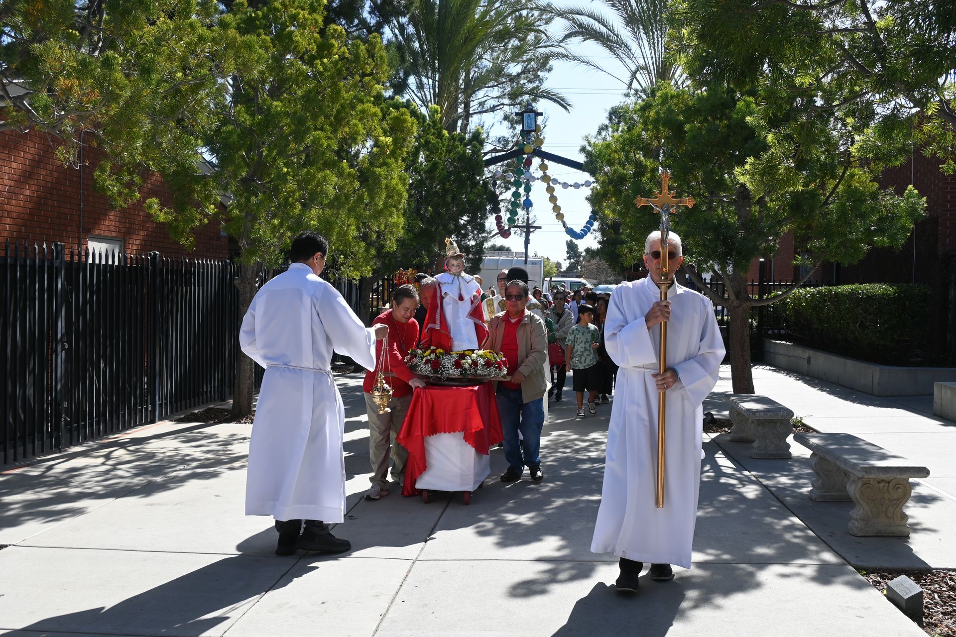 People in white robes and red garments walking outdoors on a sunny path with trees and a black fence