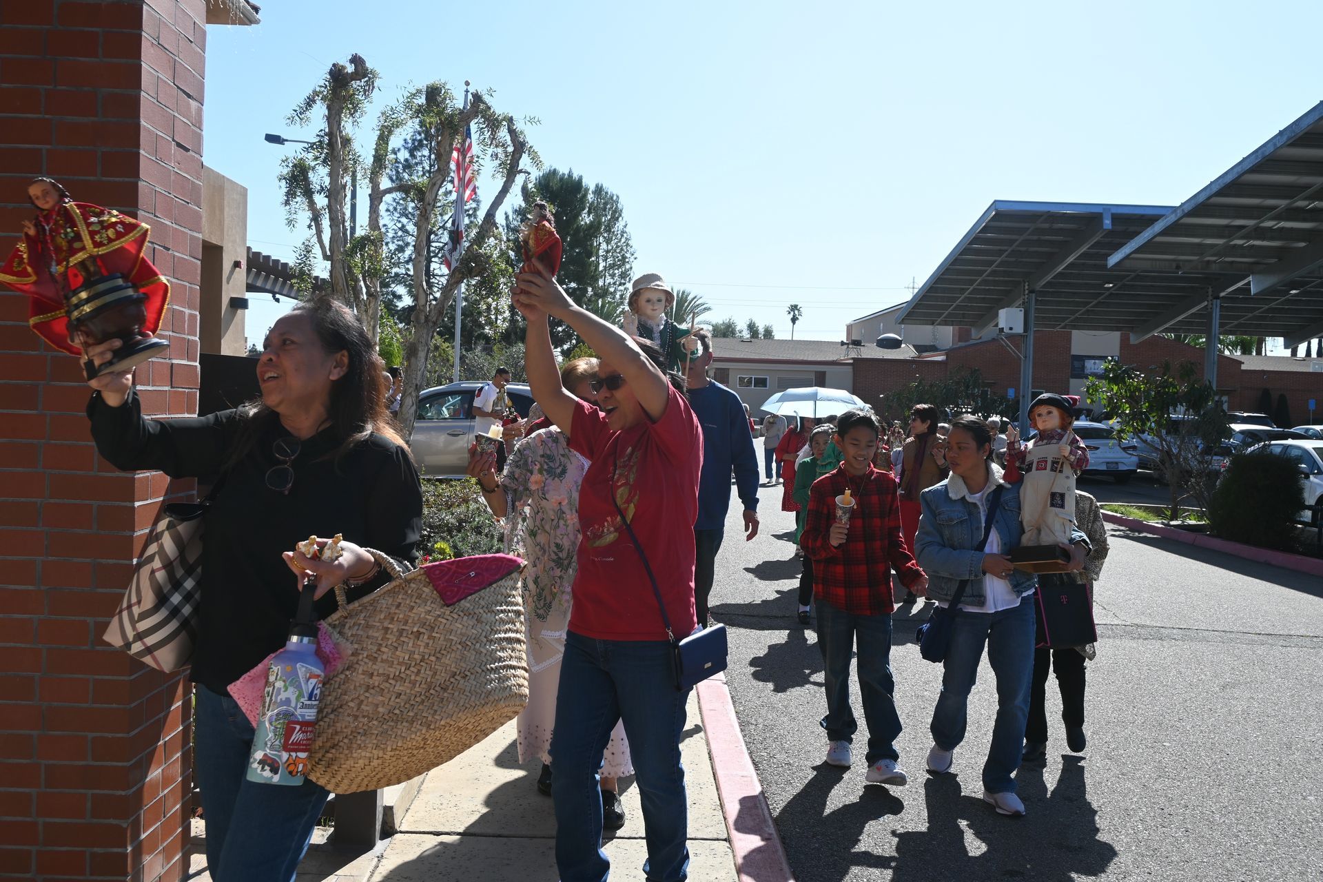 People walking outdoors near a building, some waving and holding signs in bright sunlight.