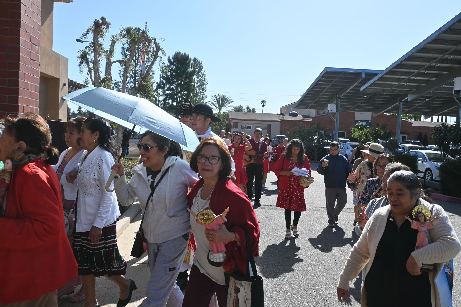 Crowd walking outdoors in bright sun, with people in colorful clothing near a building and canopy.