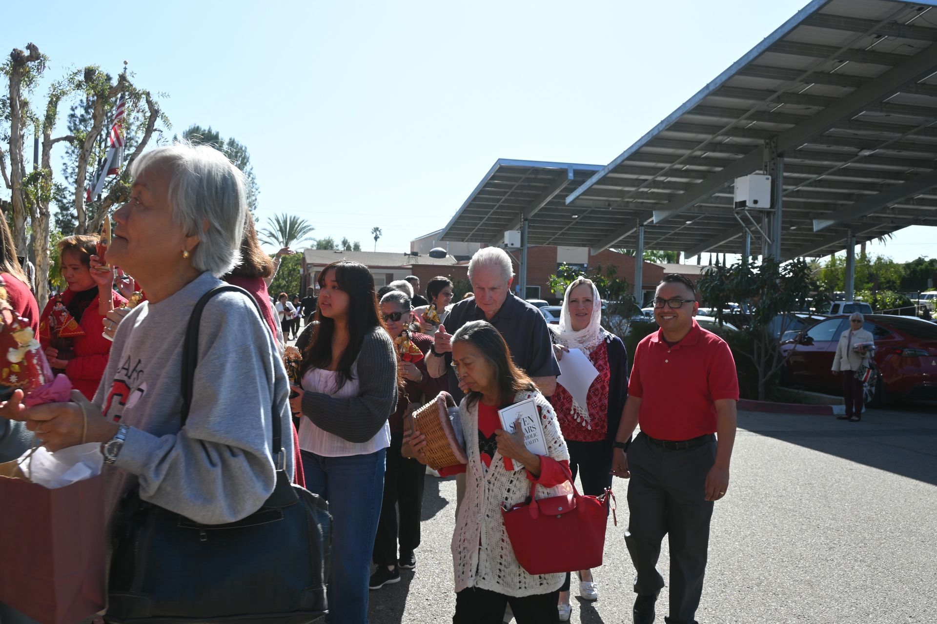 People waiting in a sunny outdoor line near a parking lot and covered structure
